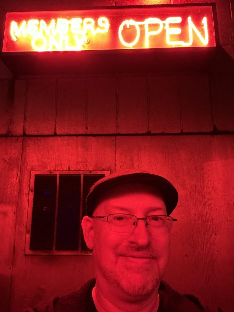 Me bathed in red light in front of a concrete wall under a red neon sign that says “OPEN MEMBERS ONLY”