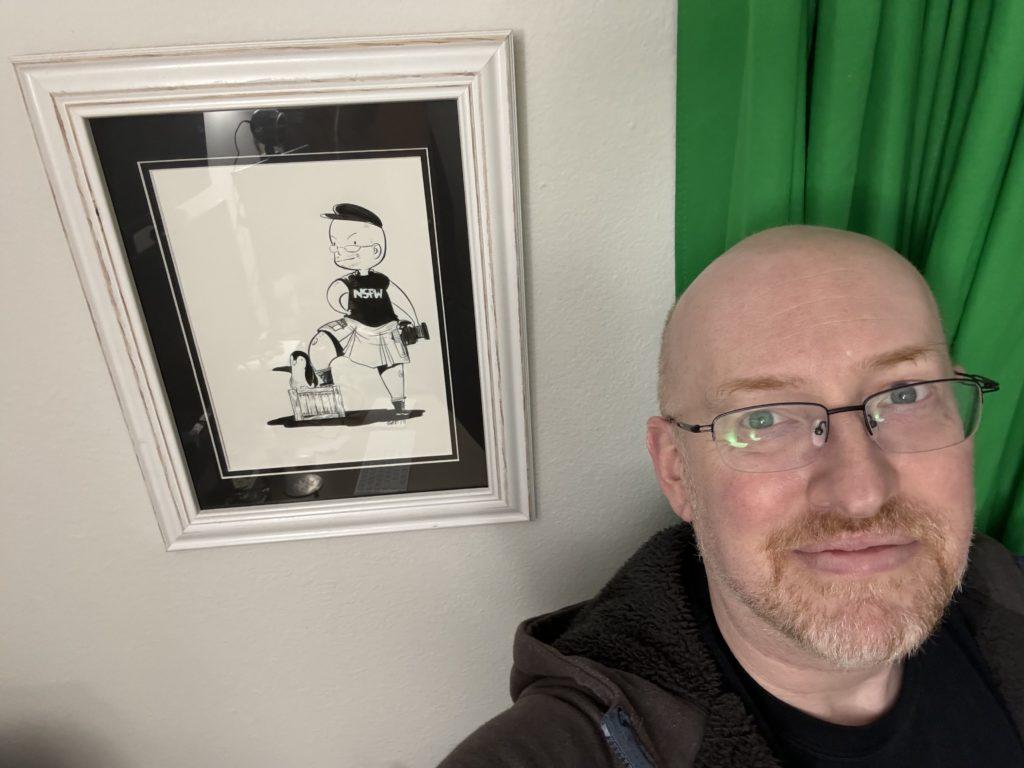 Me next to a framed piece of art of a chibi-style caricature of me holding a camera, wearing hat, kilt, and a black t-shirt that says “NSFW”, with one foot up on a wooden box with a penguin sitting next to it.