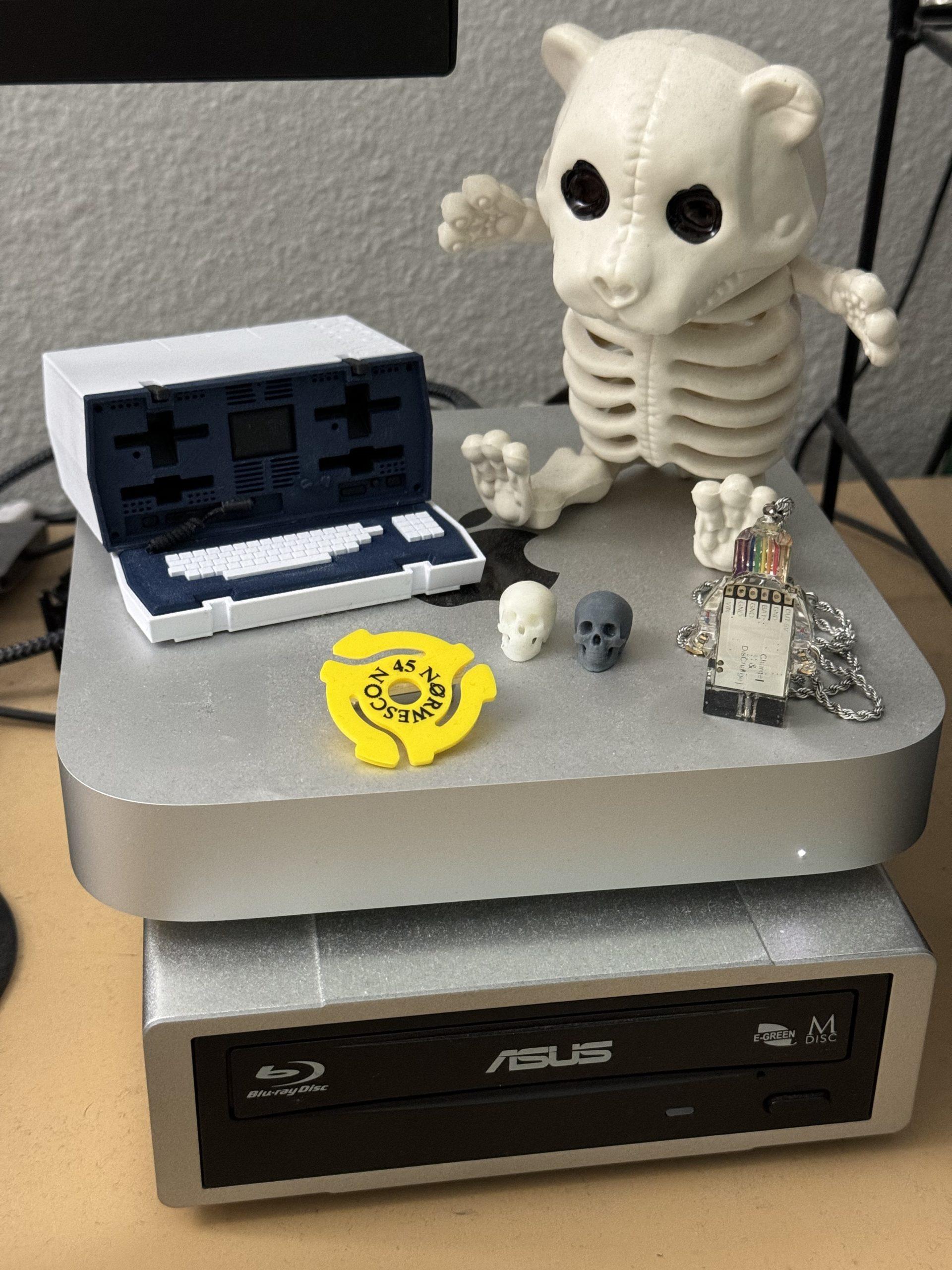 The mini Osborne sitting on top of my Mac mini, next to a teddy bear skeleton, Lego figure pendant, two tiny 3D printed skulls, and a 45 single adapter pin with the Norwescon 45 logo.