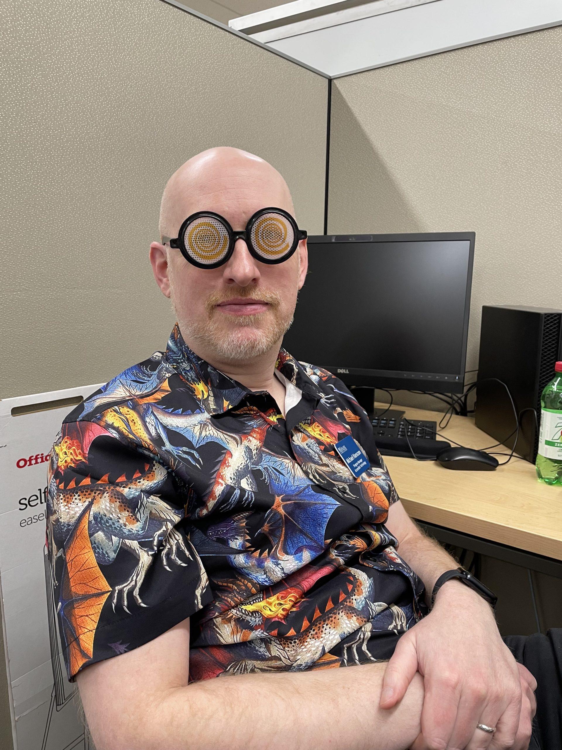 Me sitting in an office cubicle, wearing a shirt patterned with dragons, and novelty glasses that are circular with swirly plastic fake lenses.