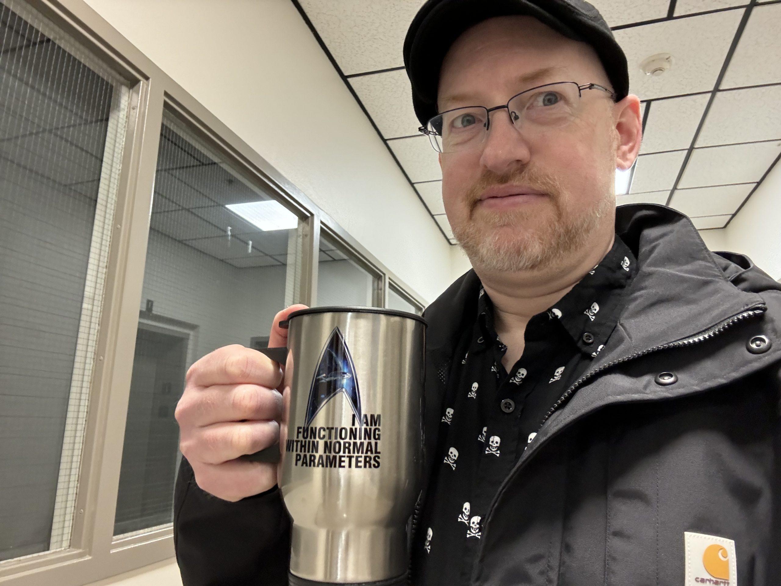 Me in a hallway wearing my winter coat, with a skeptical expression on my face, and holding a travel mug with the Starfleet delta logo and text that reads "I am functioning within normal parameters".