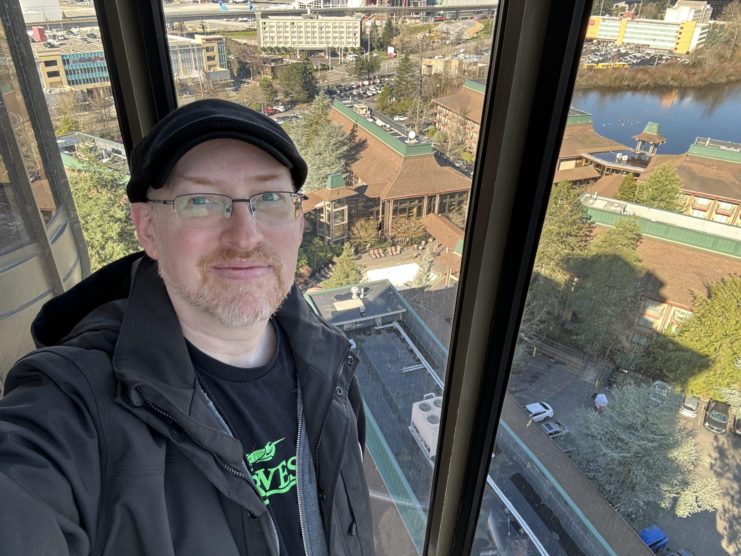 Me in a glass-walled elevator overlooking the outside of the DoubleTree Seattle Airport hotel, with several of the hotel wings and the lake visible in the distance.