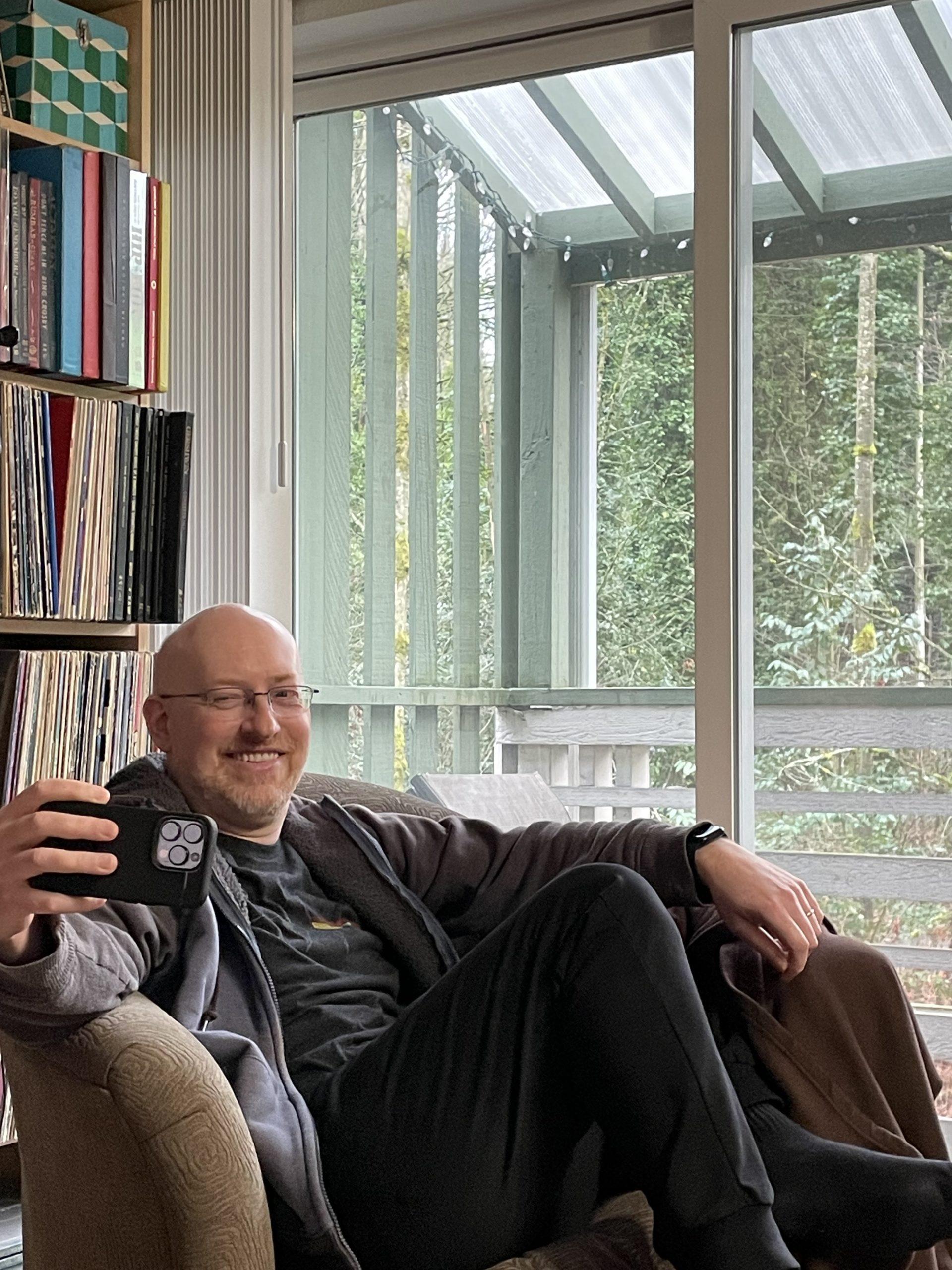 Me lounging in a chair, holding my phone out as I take a selfie, laughing as my wife takes my photo.
