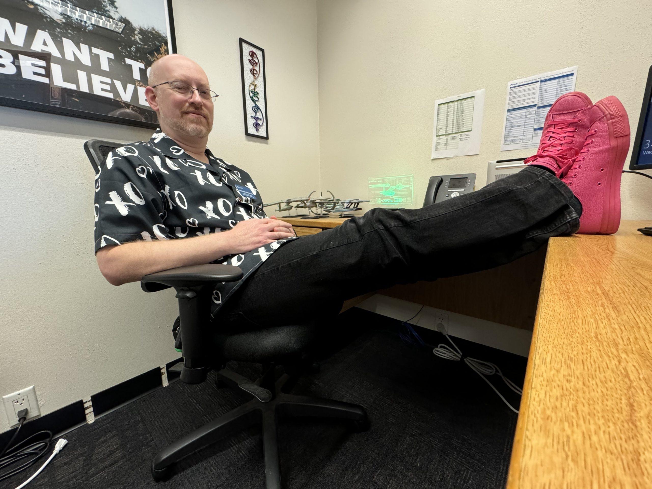 Me in my office at work, sitting in my chair with my feet up on the desk. I'm wearing a black short-sleeve button-up shirt with white X's and O's, black jeans, and bright pink Converse shoes.