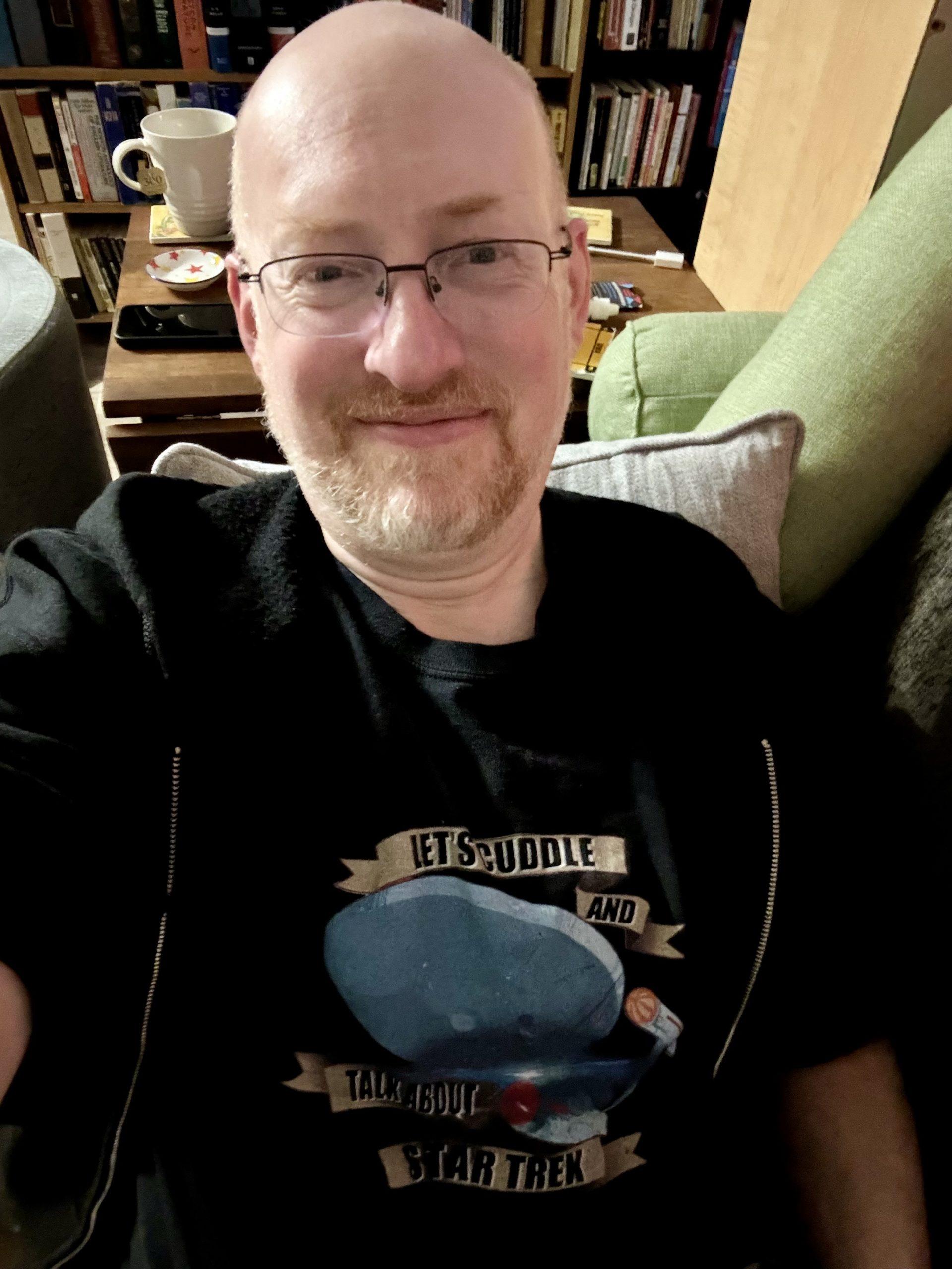 Me on our couch, wearing a shirt with an image of the TOS Enterprise and text that says 'Let's cuddle and talk about Star Trek'.