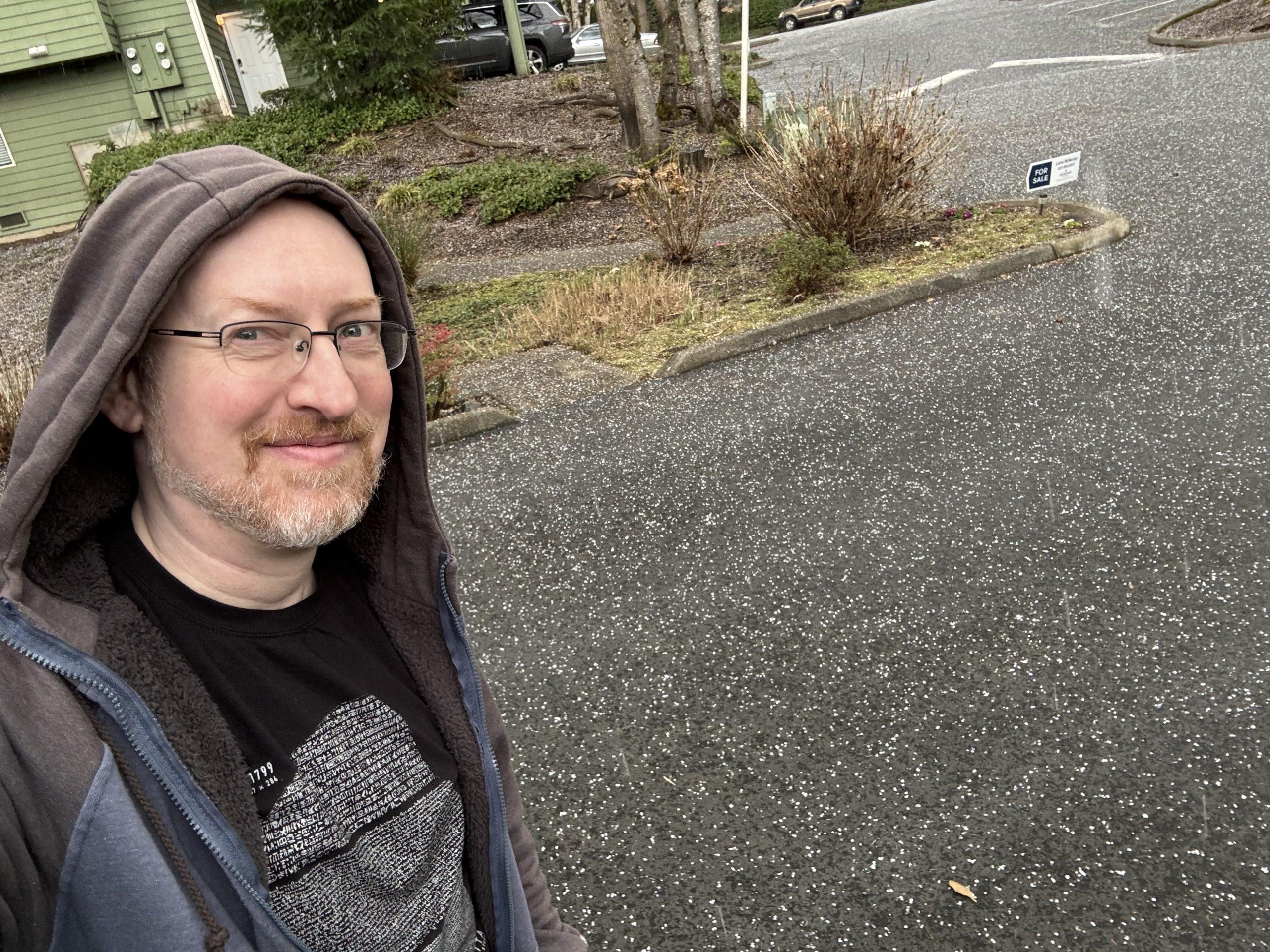 Me standing outside, with my hoodie pulled up over my head, with a very visible scattering of hail across the ground.