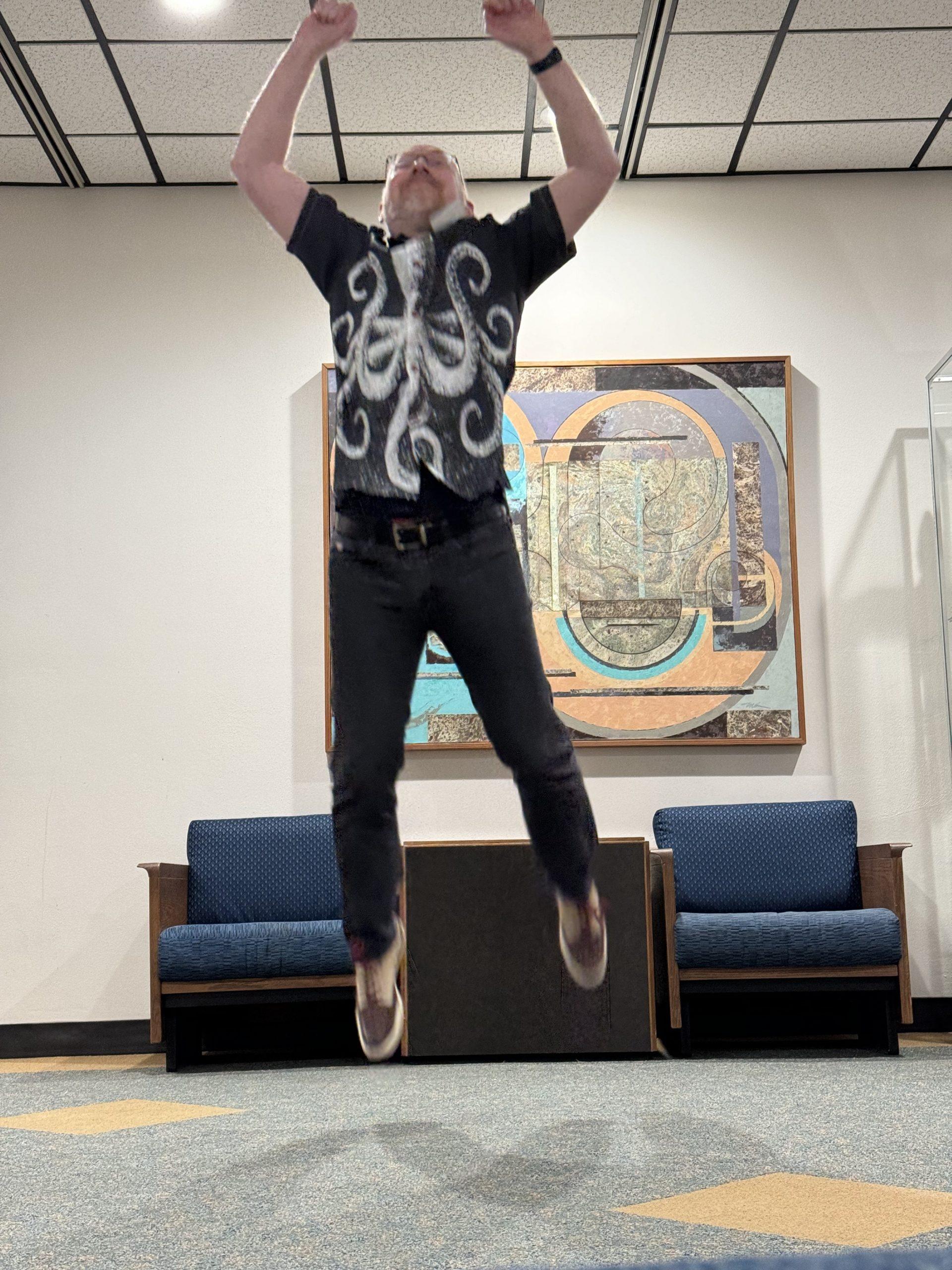 Me, wearing jeans and a shirt with an octopus print, looking quite goofy as I jump into the air with my arms raised above my head.