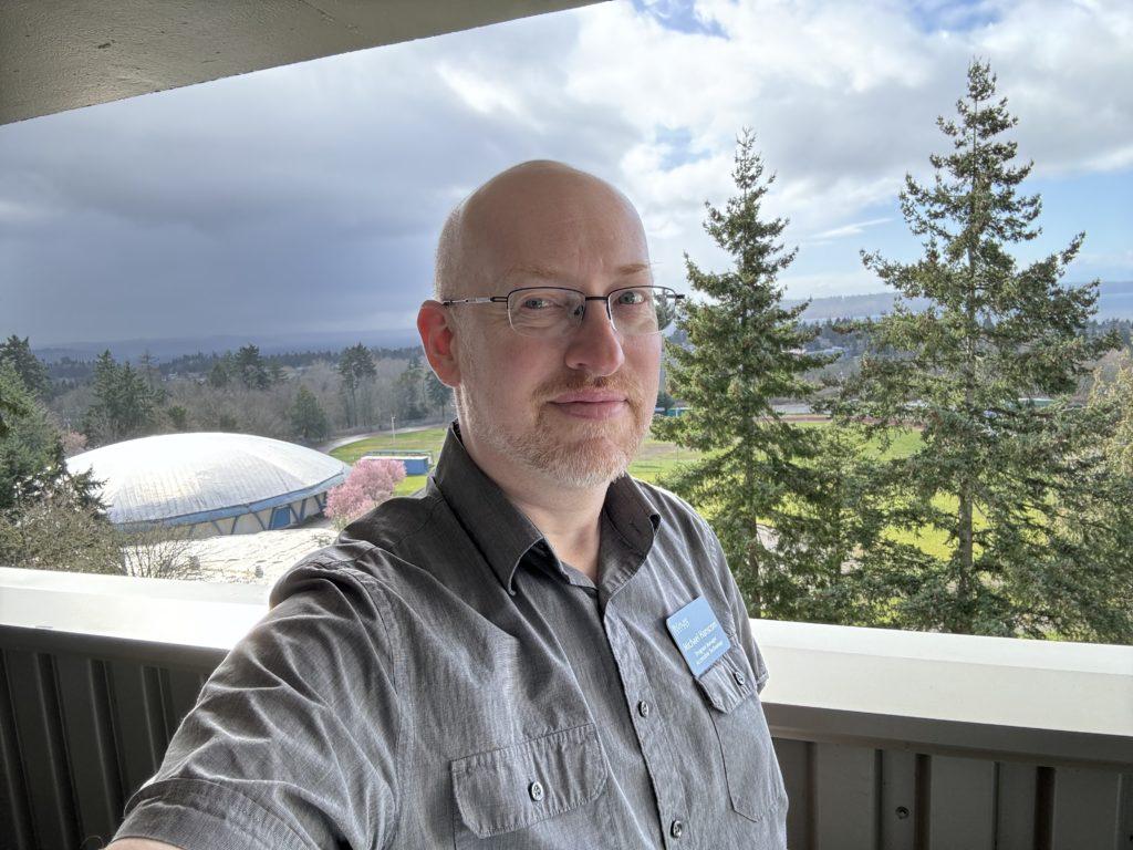 Me on my work balcony overlooking trees and sports fields, with storm clouds in half the sky and fluffy white clouds in the other half.