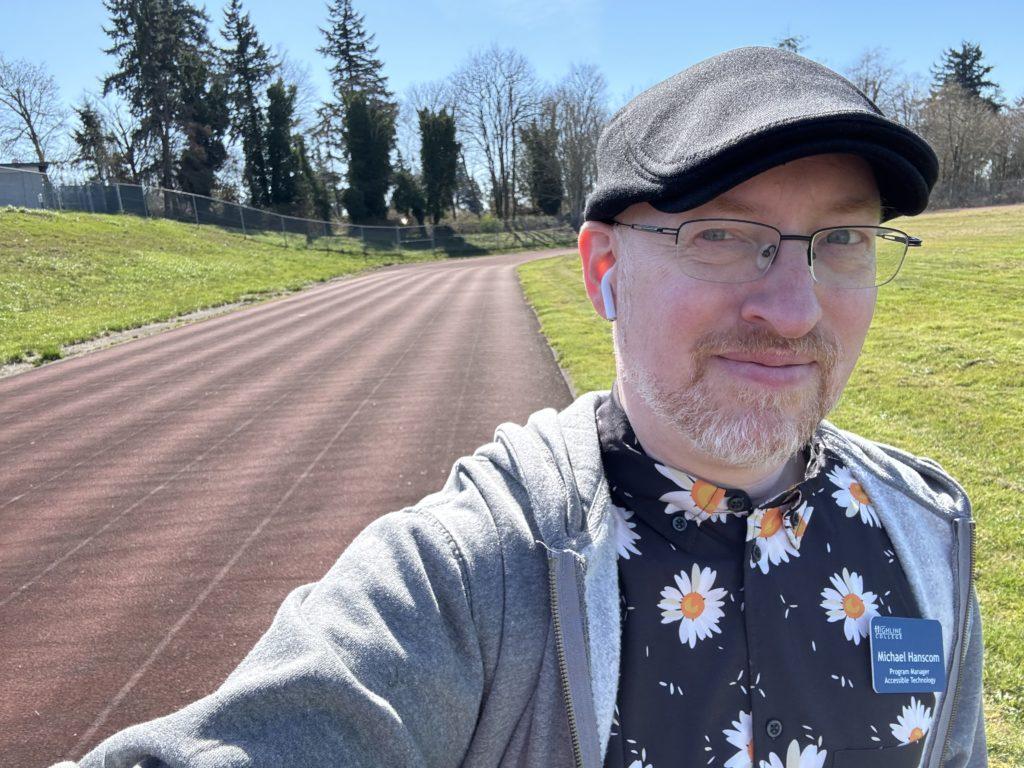 Me outside on a sunny day, walking around a sports track, wearing a light hoodie over a black shirt with daisies printed on it.