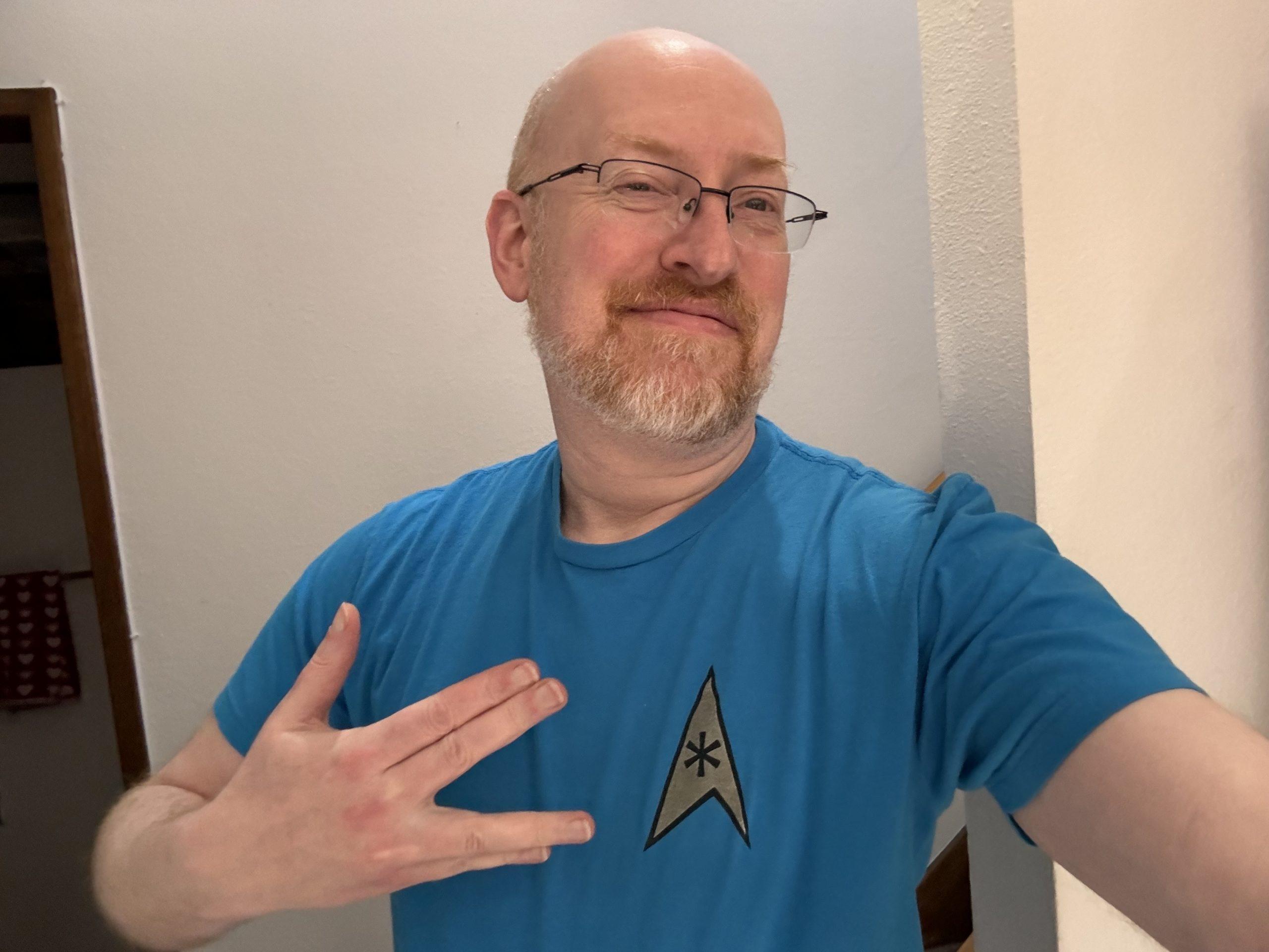 Me wearing a light blue t-shirt with a Star Tek delta shield with an asterisk. I'm making the Vulcan salute, only with my hand held sideways and with the back of my hand to the camera, and I'm kind of smirking.