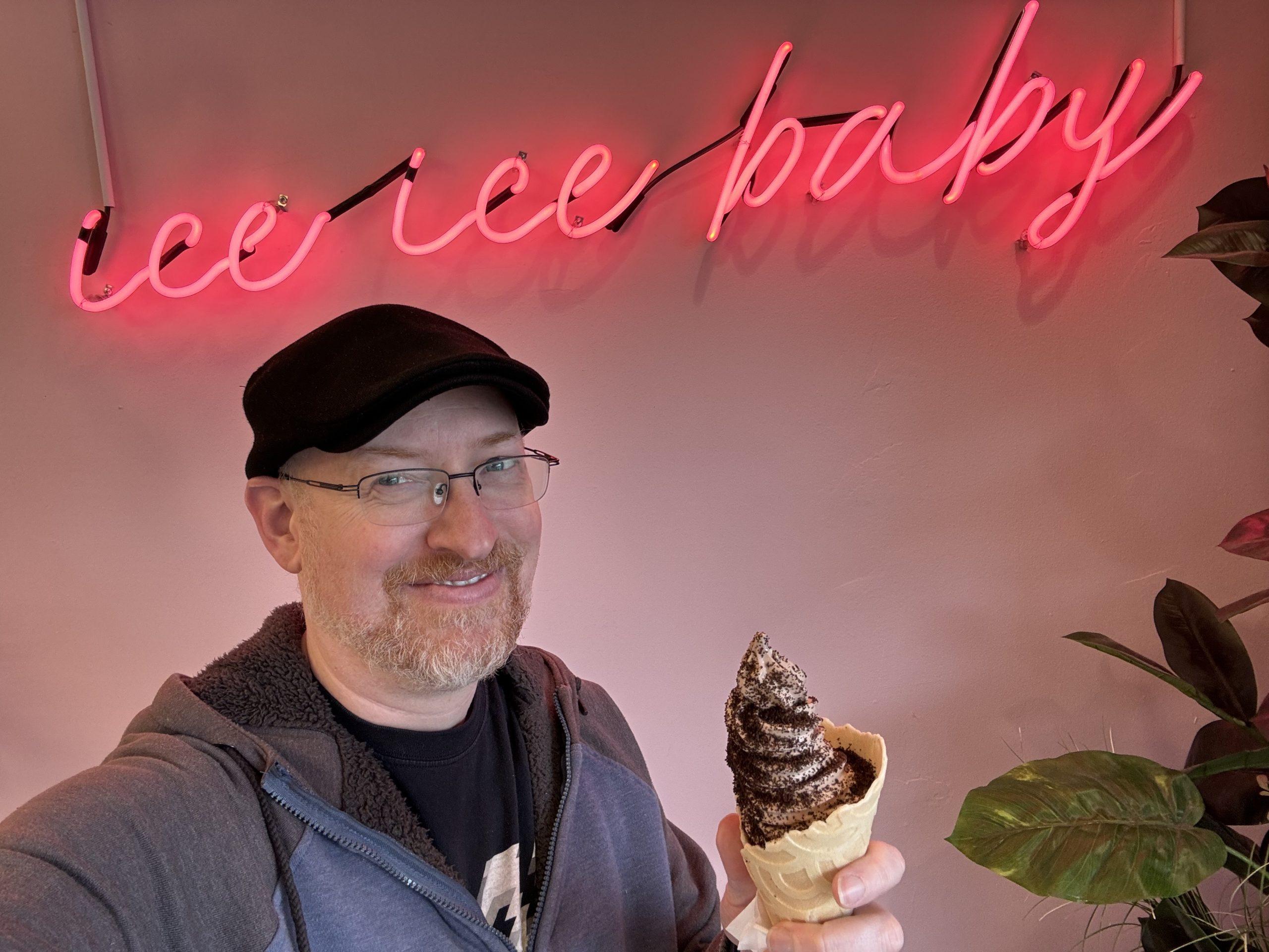 Me standing in front of a neon sign that says 'ice ice baby' while holding a waffle cone full of soft-serve ice cream.