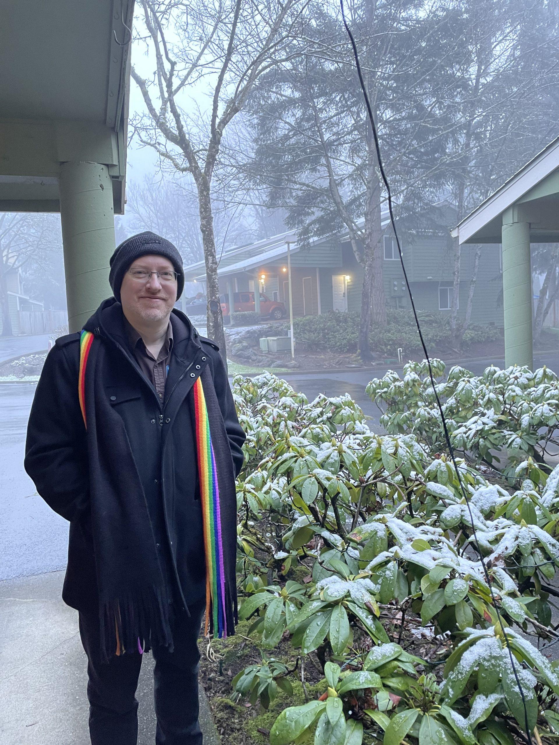 Me standing outside our house on a cold, foggy morning, wearing my winter coat, hat, and rainbow scarf, next to bushes dusted with snow.