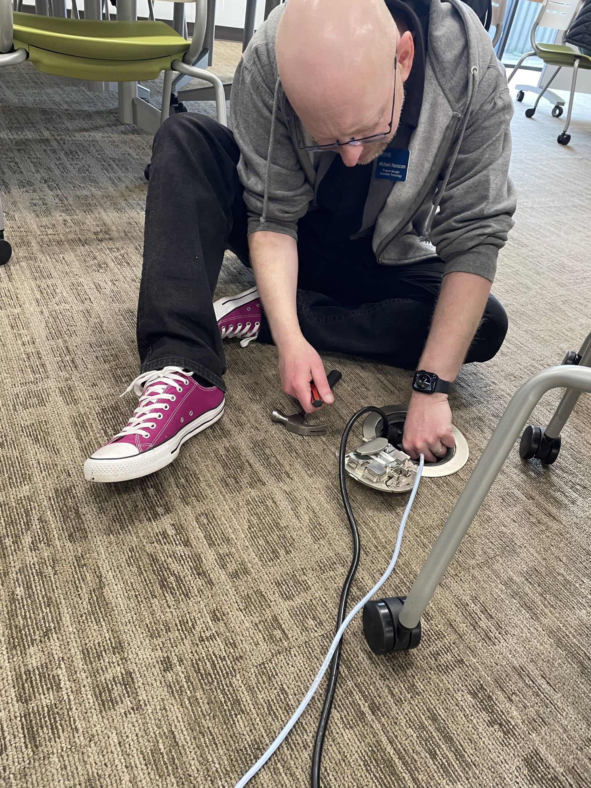 Me crouched on the floor of a classroom, holding a screwdriver in one hand as I reach into a power and network placement on the floor. A power cable and a network cable disappear into the placement.