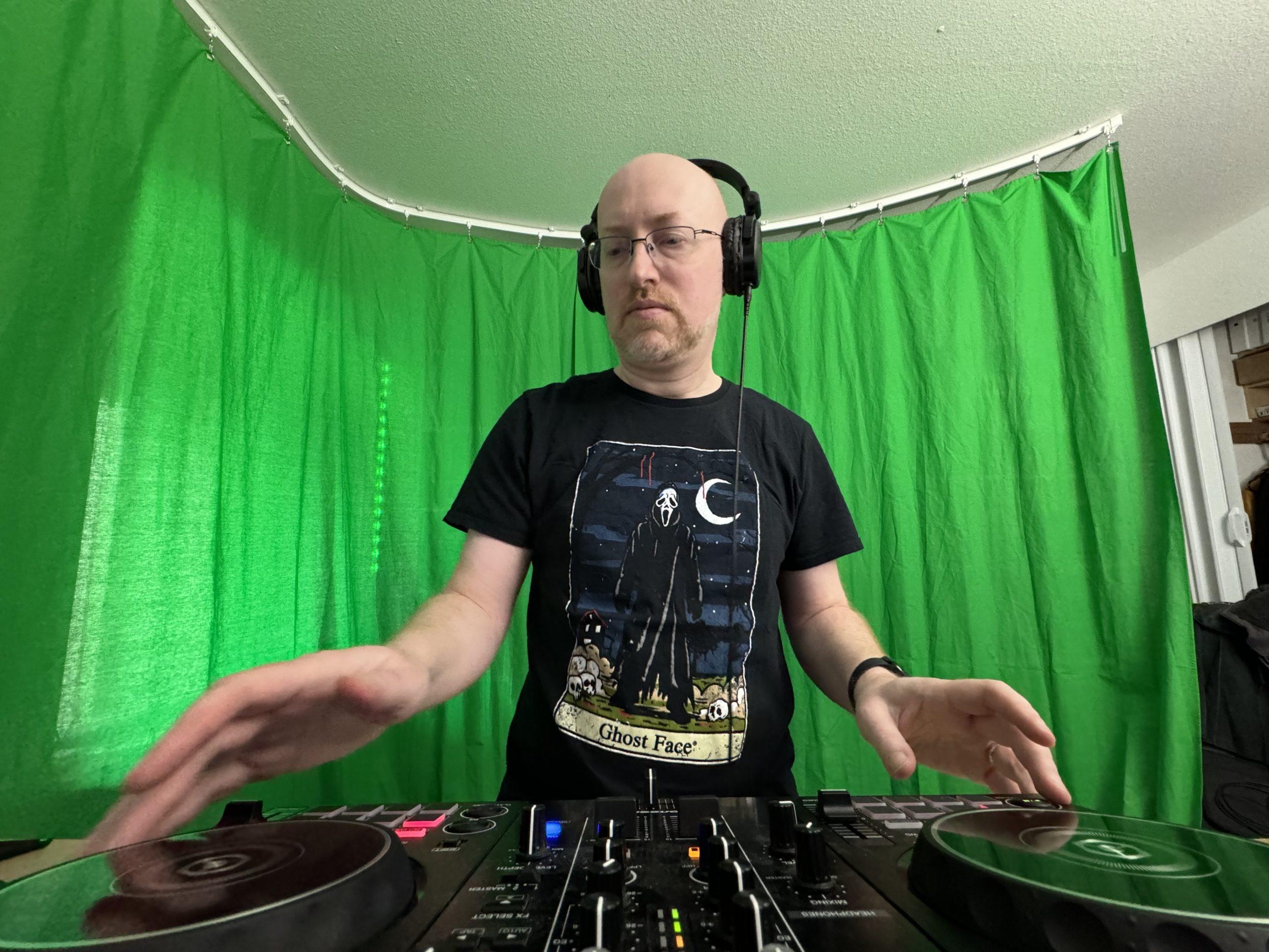 A wide-angle shot with a DJ mixer in the foreground, me wearing headphones and with my hands slightly blurry as I spin one of the mixer controllers, and a greenscreen hanging from a ceiling-mounted track behind me.