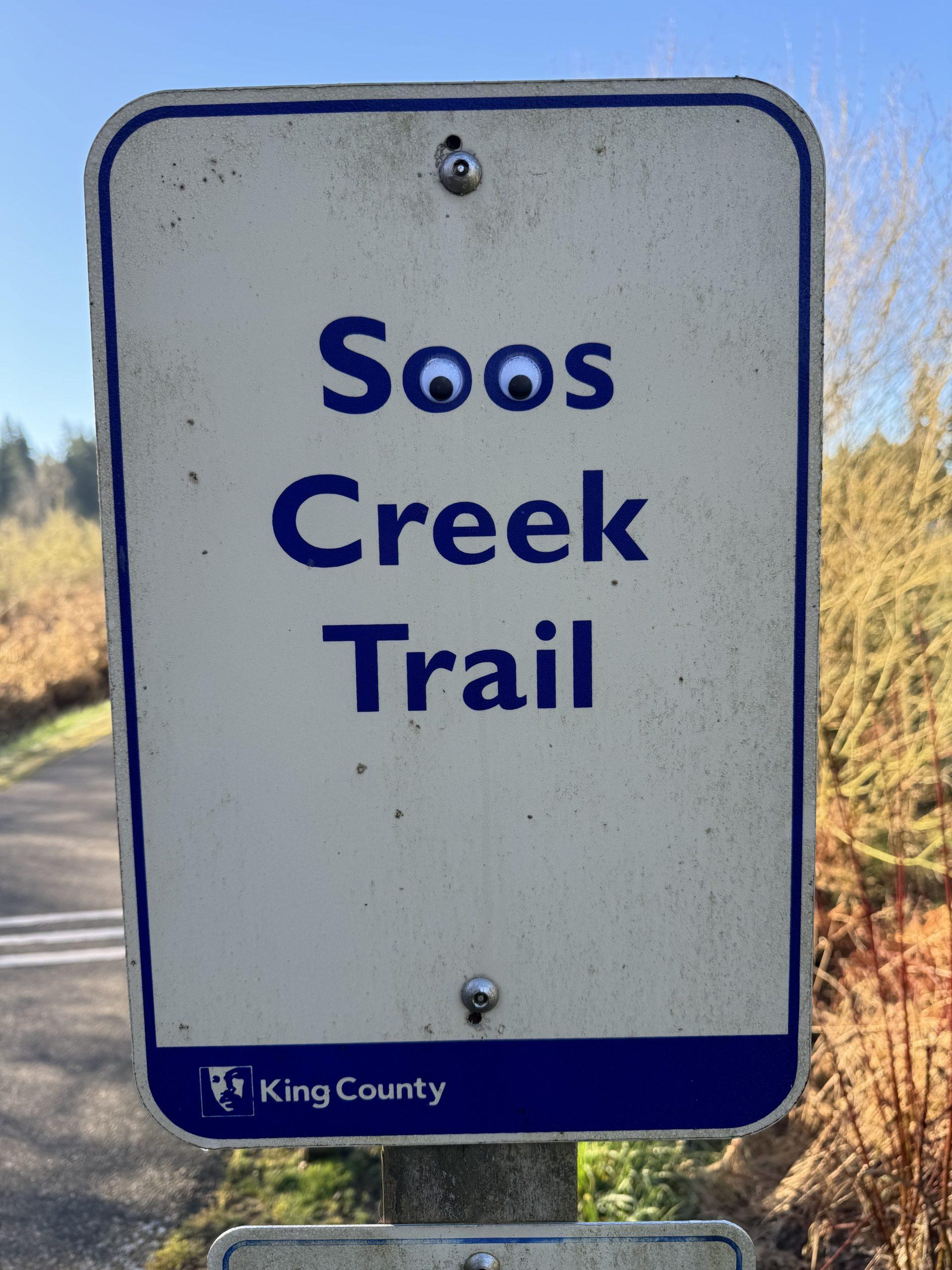 A trail sign for the Soos Creek Trail with googly eyes placed on both of the Os.