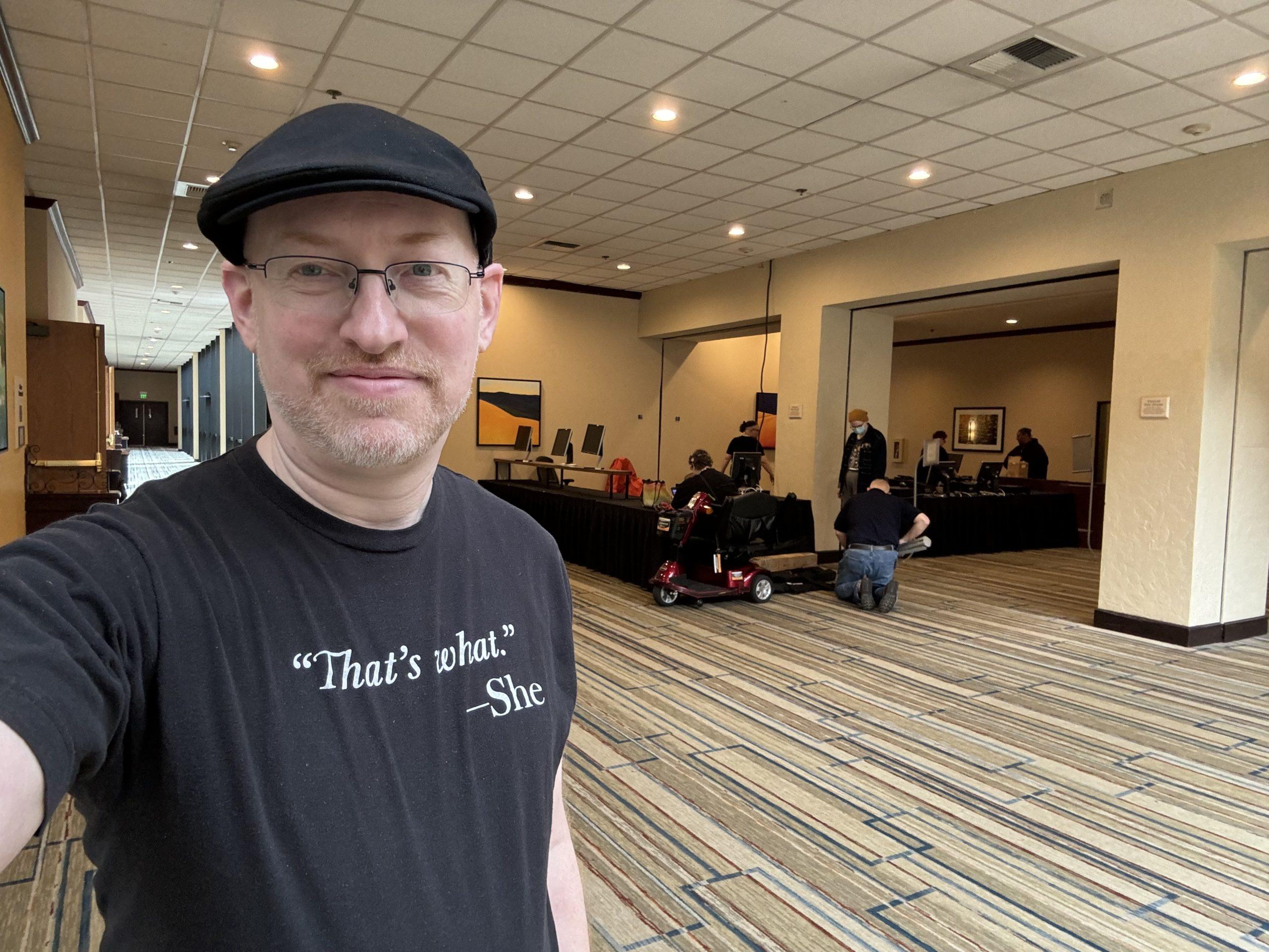 Me in a hotel lobby wearing a shirt that says, "'That's what.' She". Behind me are a few people setting up tables.