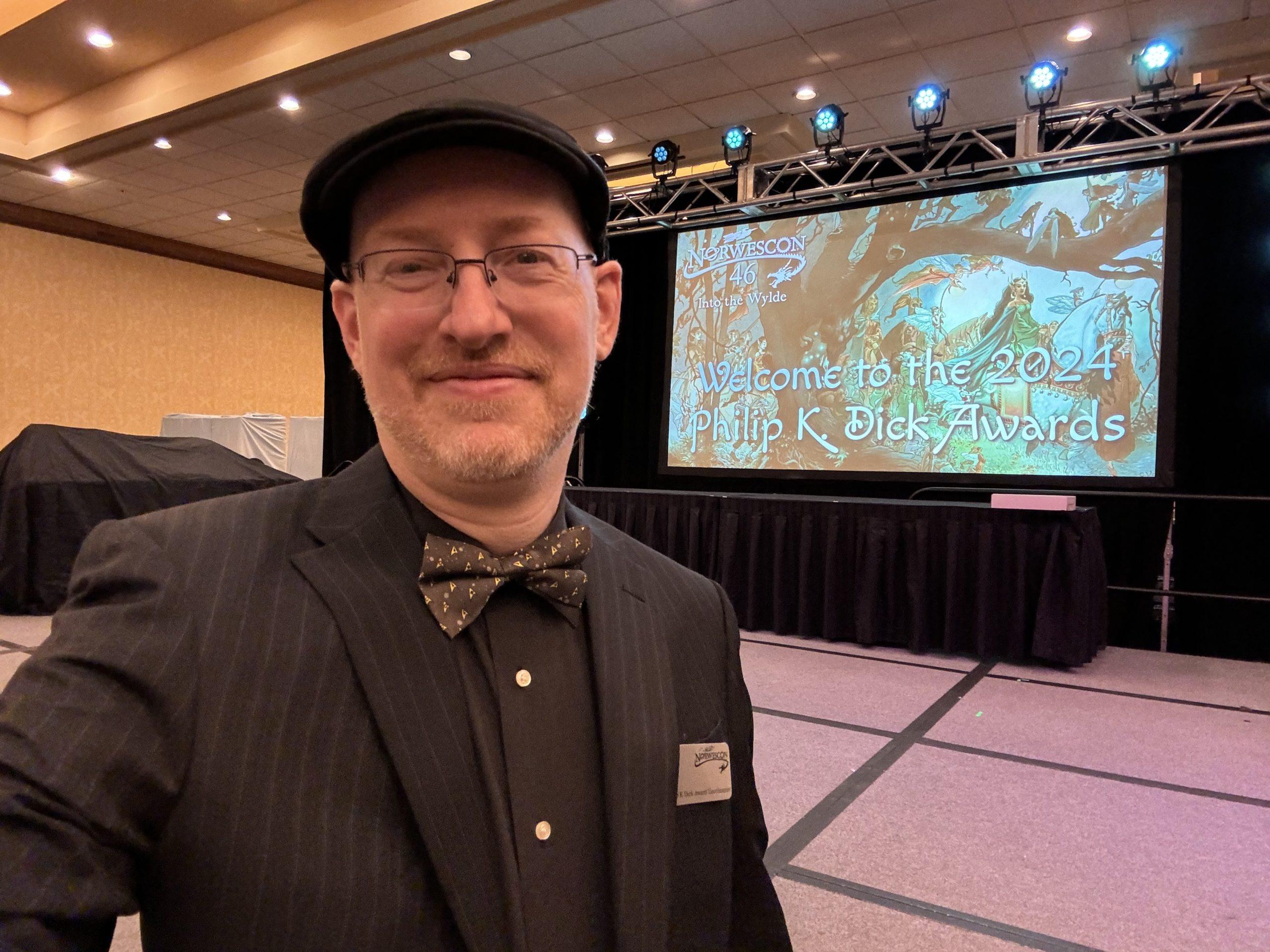 Me wearing a black cap, black suit jacket with grey pinstripes, black shirt, and Star Trek bow tie, in front of a stage with a screen showing a slide that says 'Welcome to the 2024 Philip K Dick Awards'