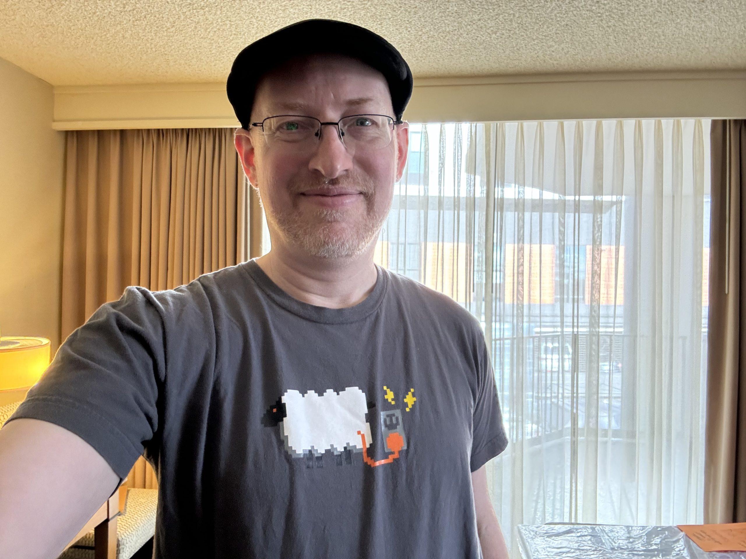 Me in my hotel room wearing a grey t-shirt with pixel art of a fluffy white sheet plugged into an electric outlet with an orange cord.