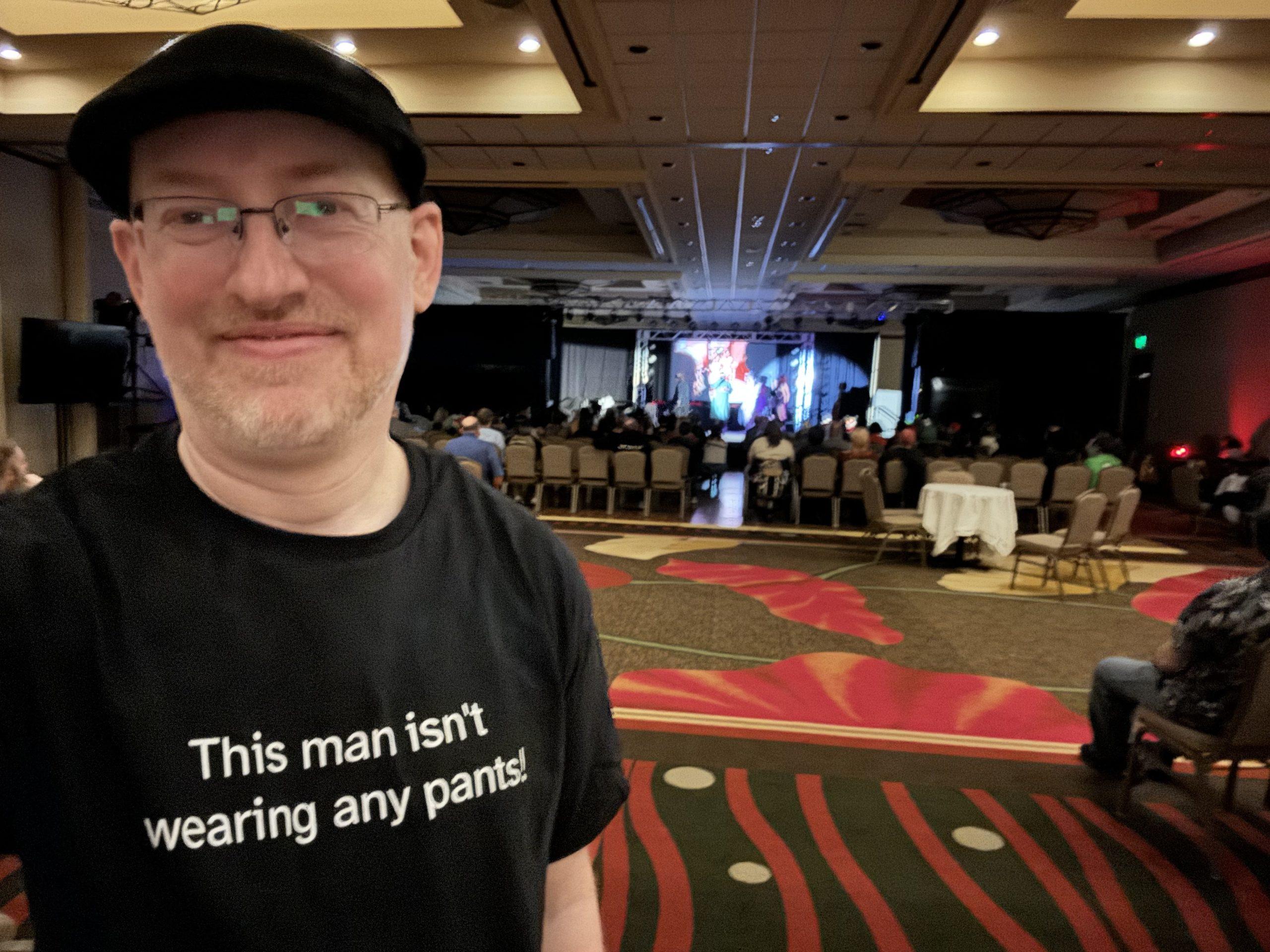 Me in the back of a hotel ballroom, with a crowd watching the Rocky Horror Picture Show in the background; I'm wearing a black t-shirt with white text that says 'This man isn't wearing any pants!'