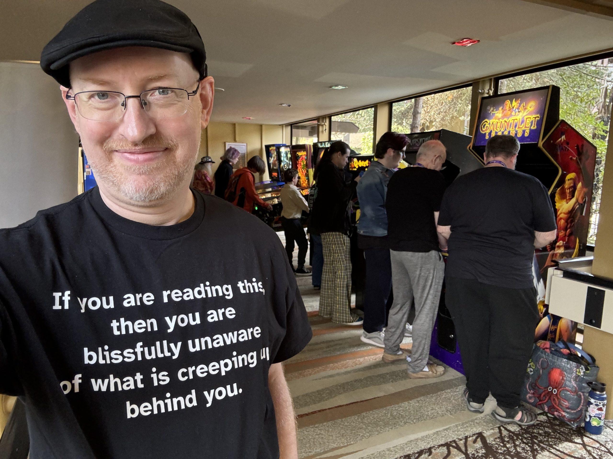 Me in front of a very busy lineup of pinball and arcade machines, wearing a shirt that says, 'if you are reading this, then you are blissfullly unaware of what is creeping up behind you'.