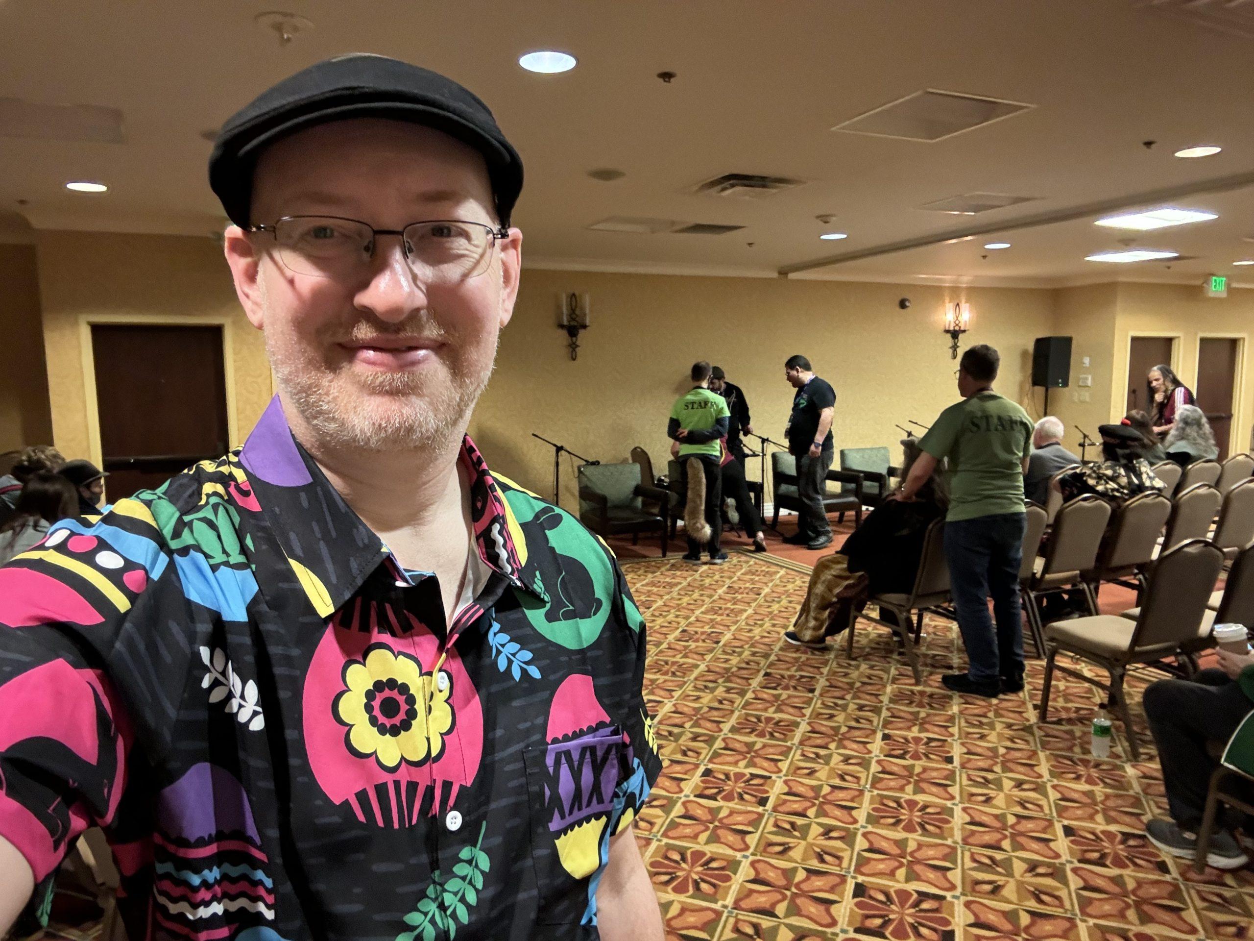 Me in a hotel meeting room with a few people around chairs behind me; I'm wearing a black button-up short-sleeve shirt with large colorful Easter eggs on it.