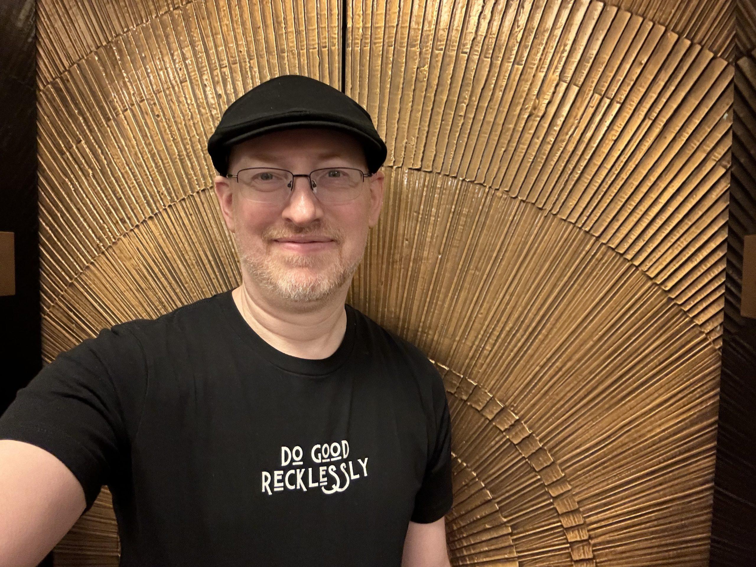 Me in front of gold patterned elevator doors, wearing a t-shirt that says 'do good recklessly'.