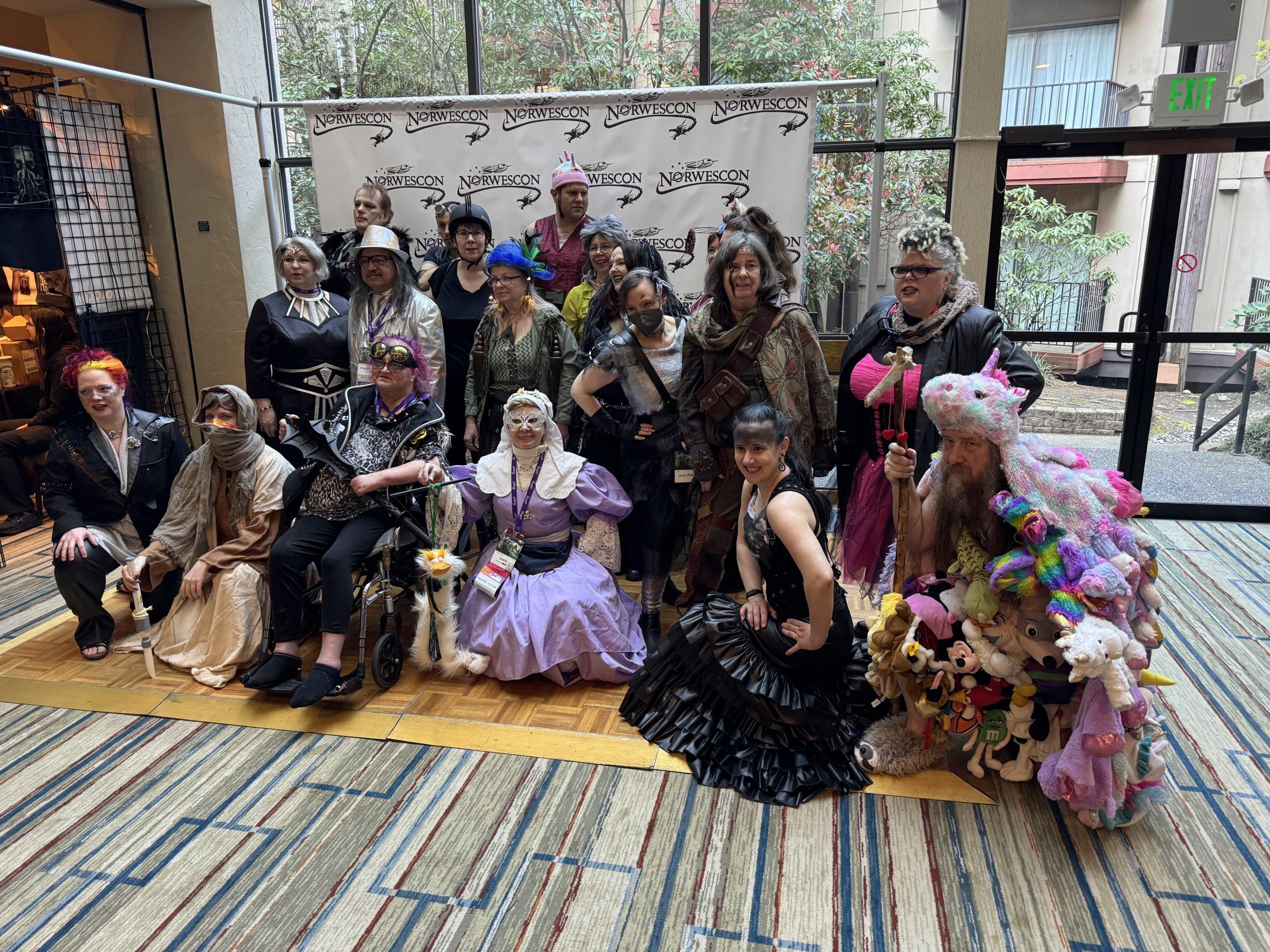 A group of people in front of a photo backdrop with the Norwescon logo, dressed in outfits inspired by the idea of a Mad Max-themed prom, mashing distressed clothing with formal wear.