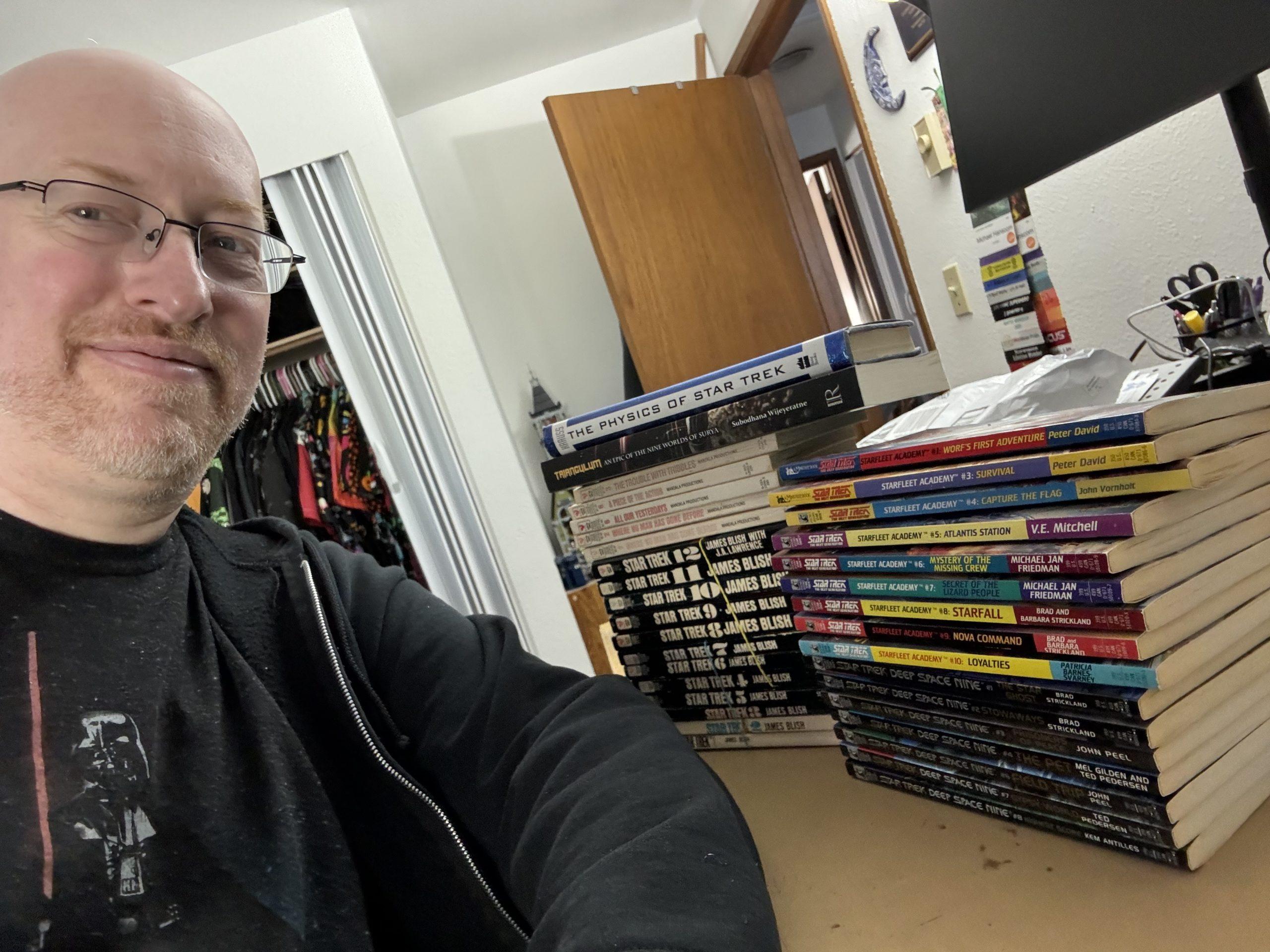 Me at my desk at home, next to two stacks of Star Trek books, including all the James Blish logs, some of the photonovels, and near complete sets of the TNG and DS9 YA series.