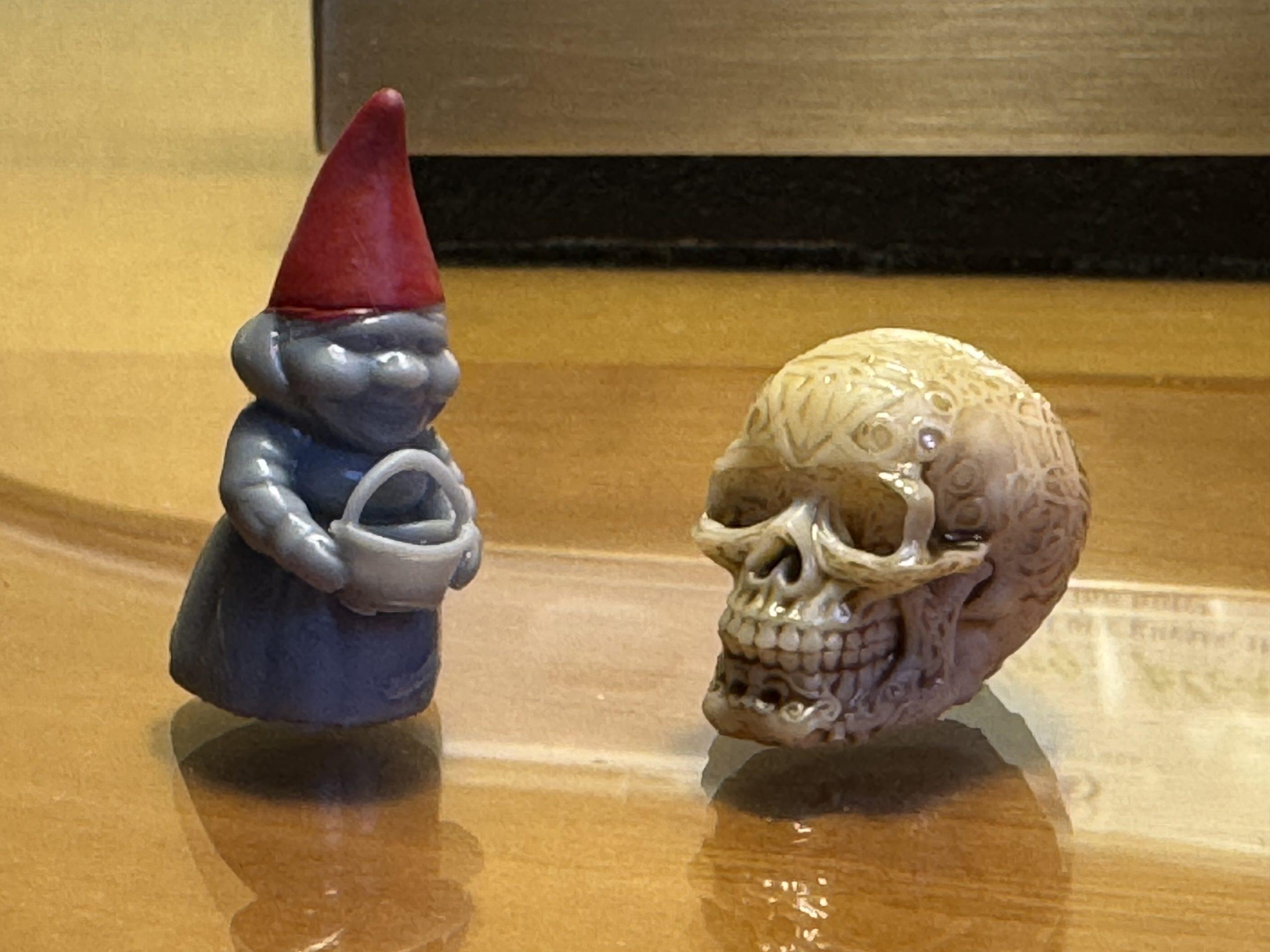 A tiny female gnome printed in grey with a painted red hat, and a tiny ornately carved skull printed in ivory with tinted carvings.