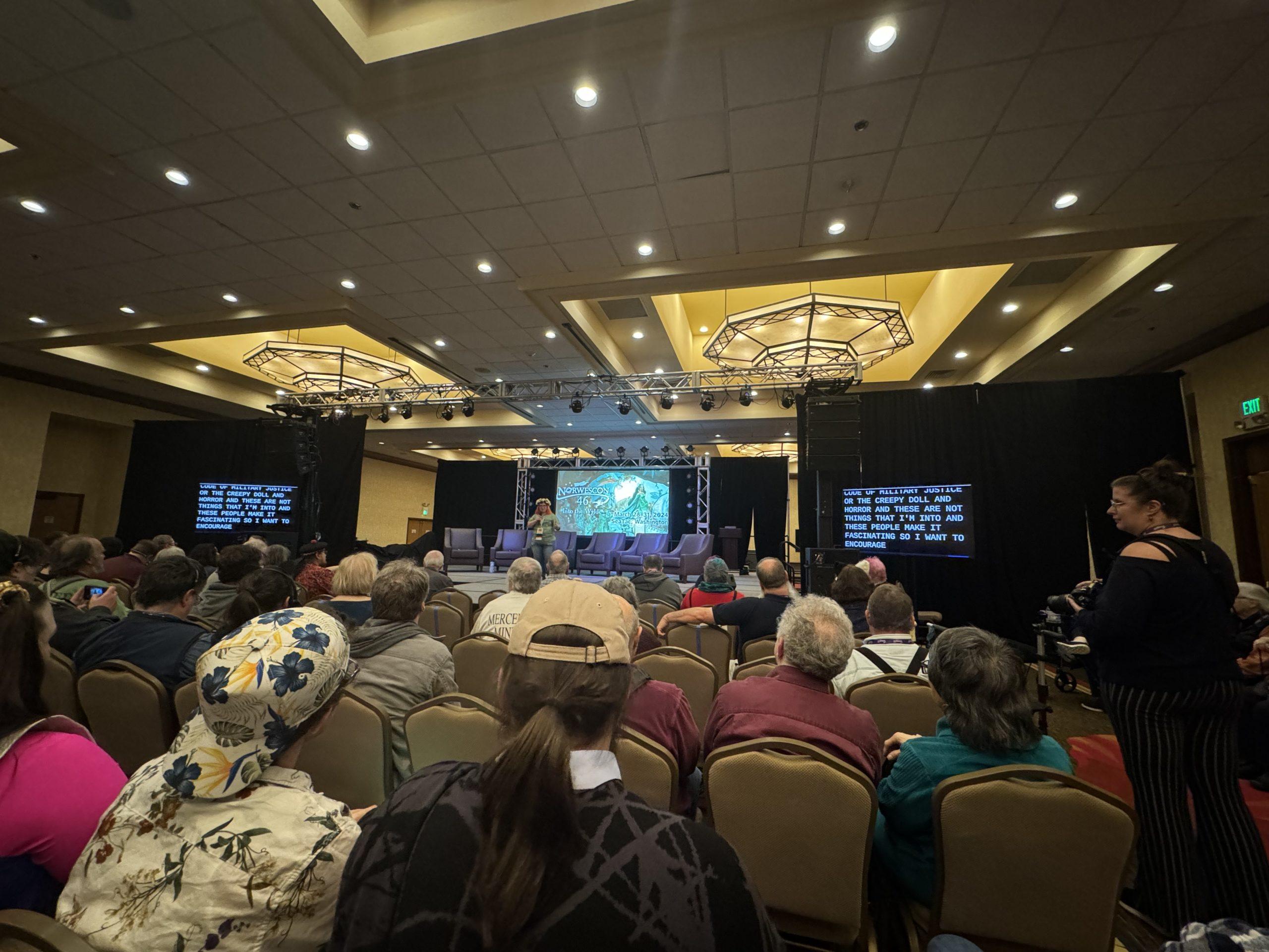 A hotel ballroom with a stage set up at one end and an audience sitting in chairs facing the stage; one person and five empty chairs are on the stage in front of a large screen with the convention artwork projected on it; large video displays on either side of the stage show captions.