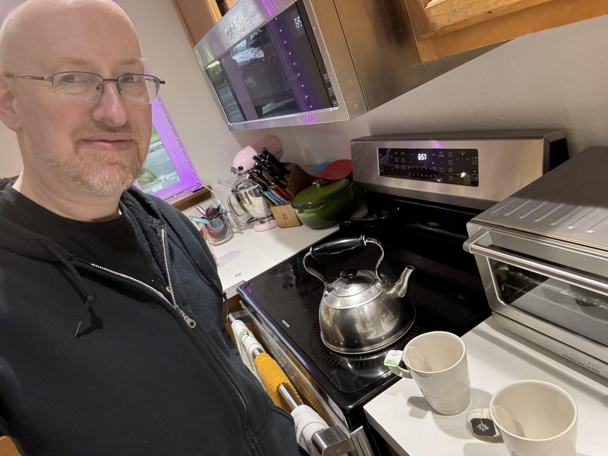 Me standing at the range in our kitchen. There is a teakettle on the range, and two mugs with teabags sitting on the kitchen counter.