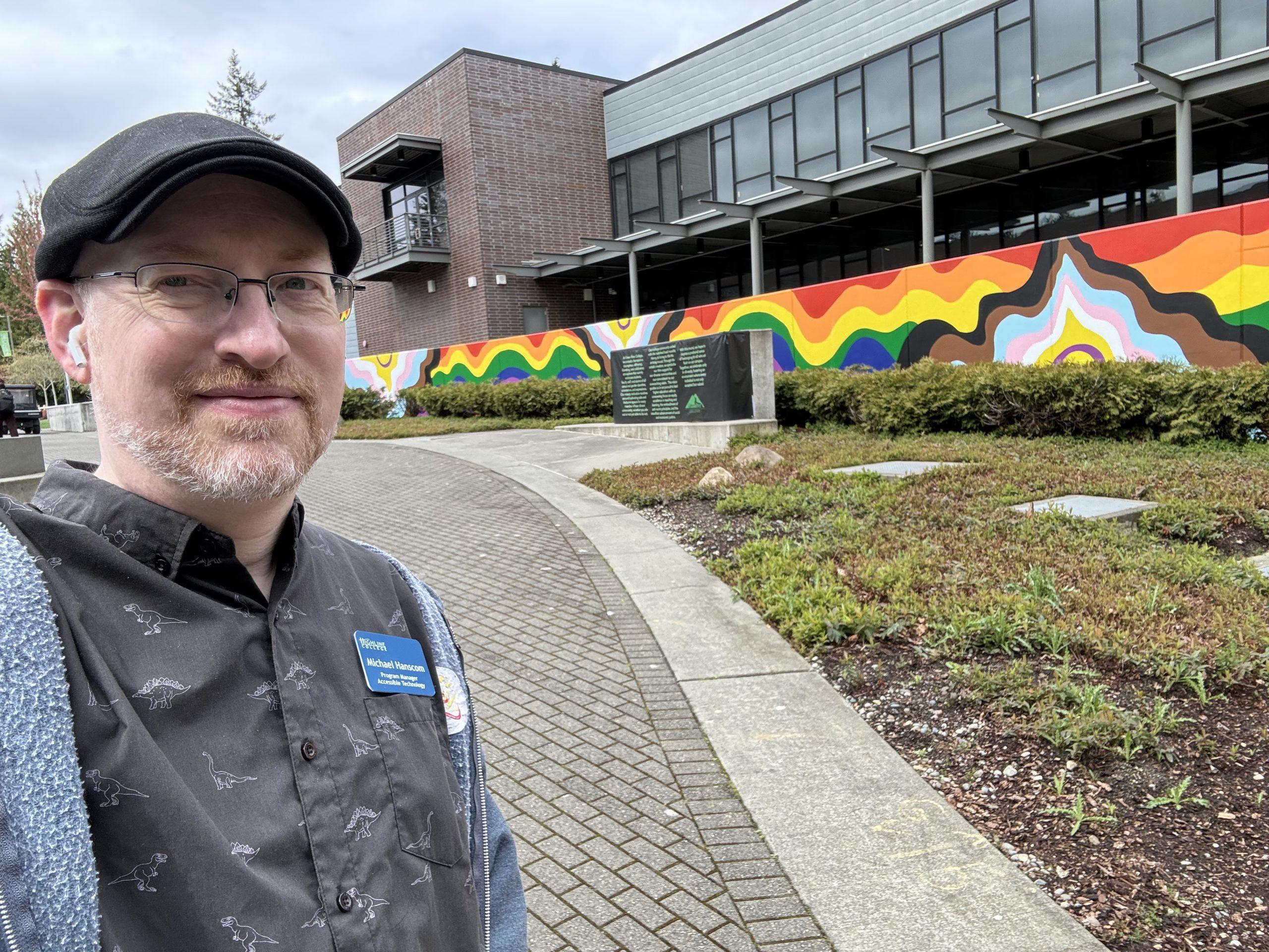 Me outside the Green River College student union building, in front of a long mural of Pride colors, including trans and black and brown pride stripes.