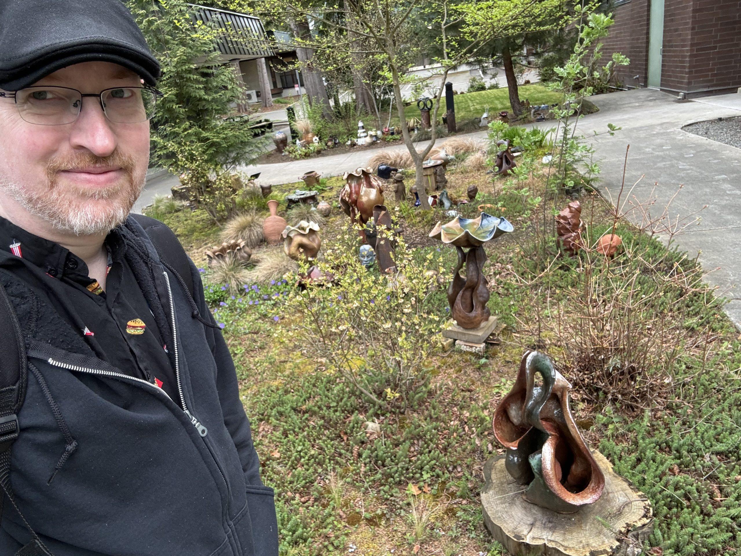 Me standing in front of a landscaped area decorated with various bits of pottery. There are vases, a bird bath, and several abstract forms. More pottery is visible in the background across a paved path.