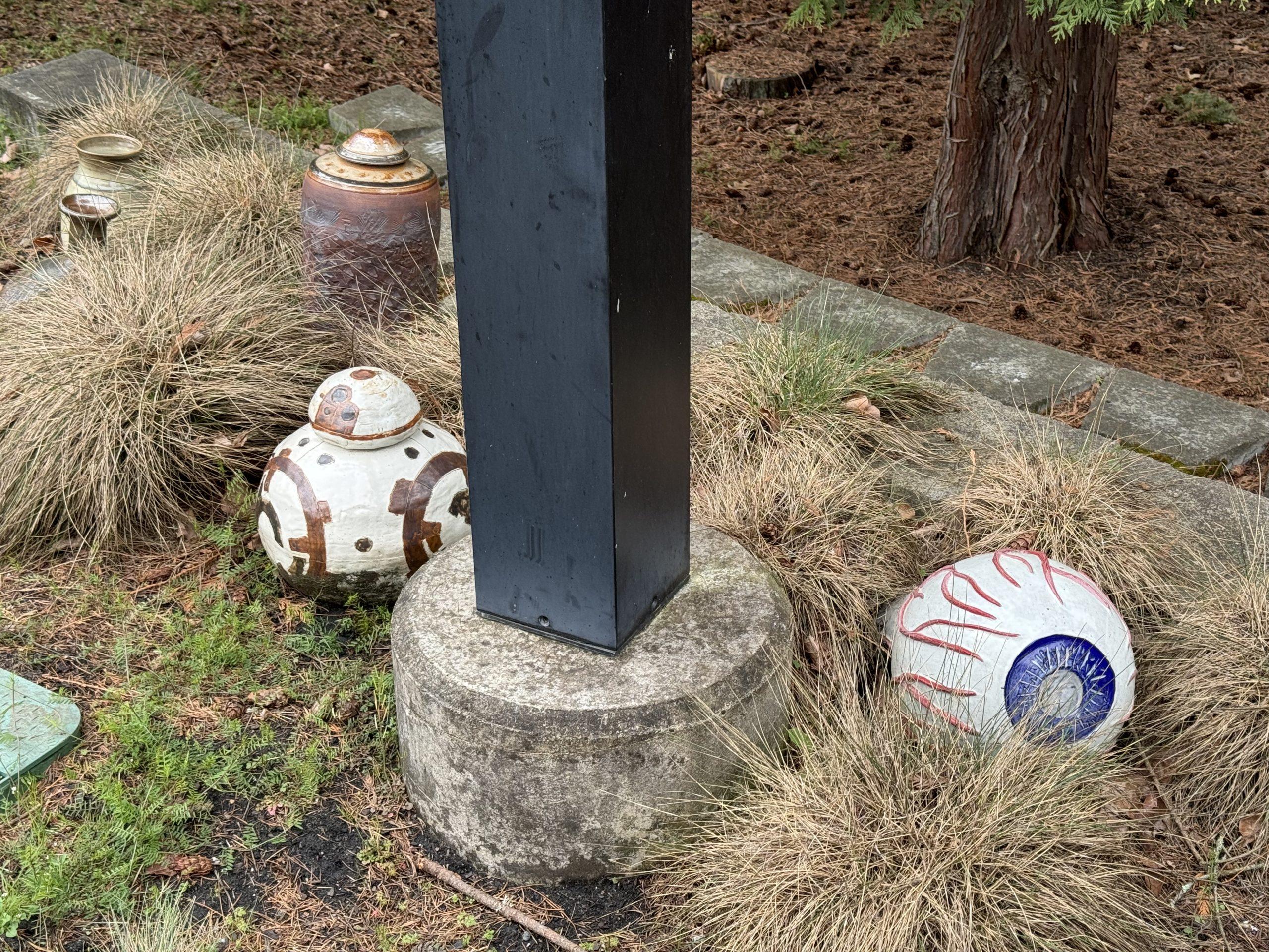 Pottery sculptures tucked into grasses near the base of a light pole. One looks like an eyeball, another like the BB-8 droid from Star Wars, with vases and urns behind them.