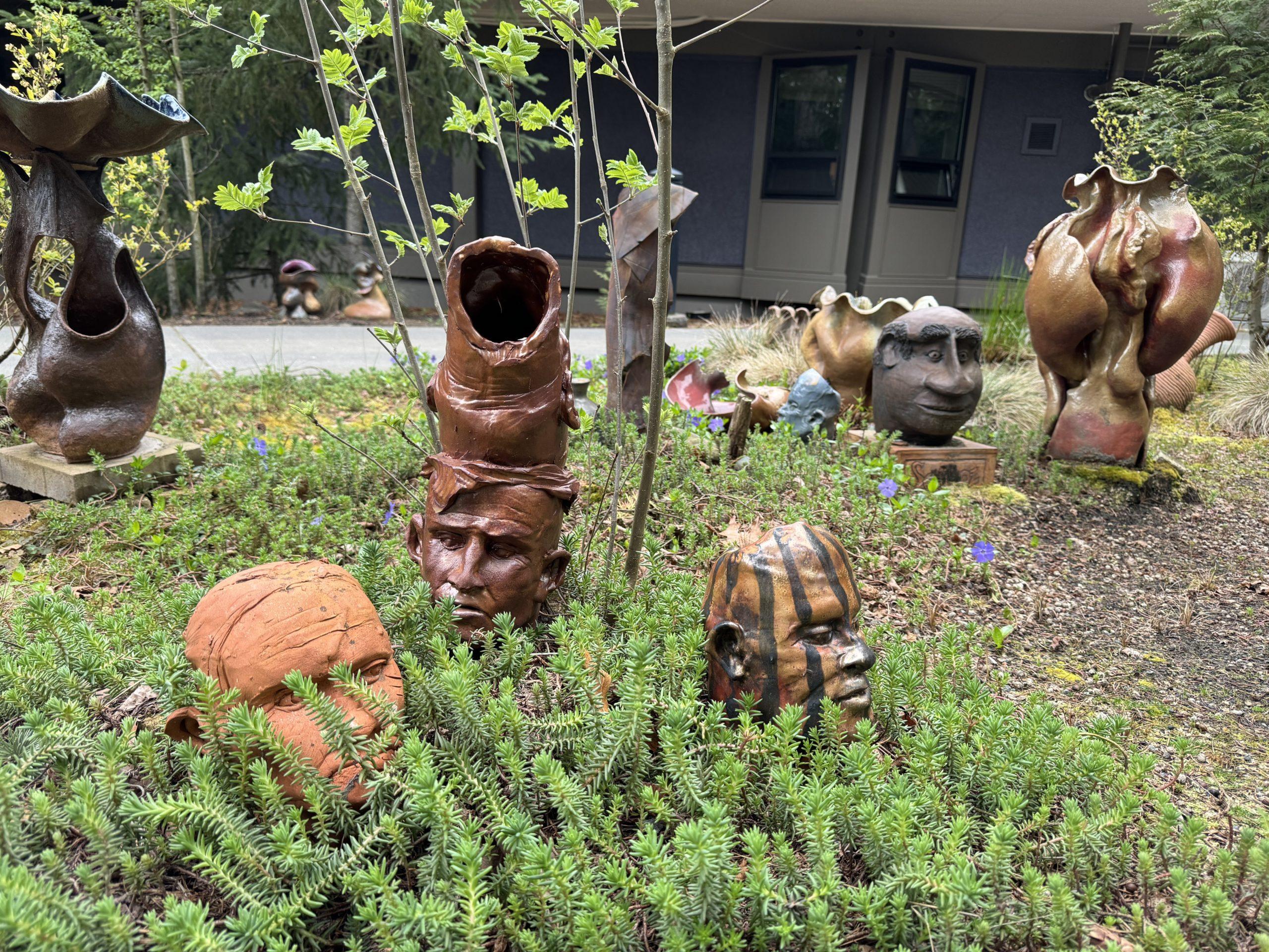 Several pottery pieces tucked in among low bushes, most of them sculpted heads, with other more abstract forms behind them.