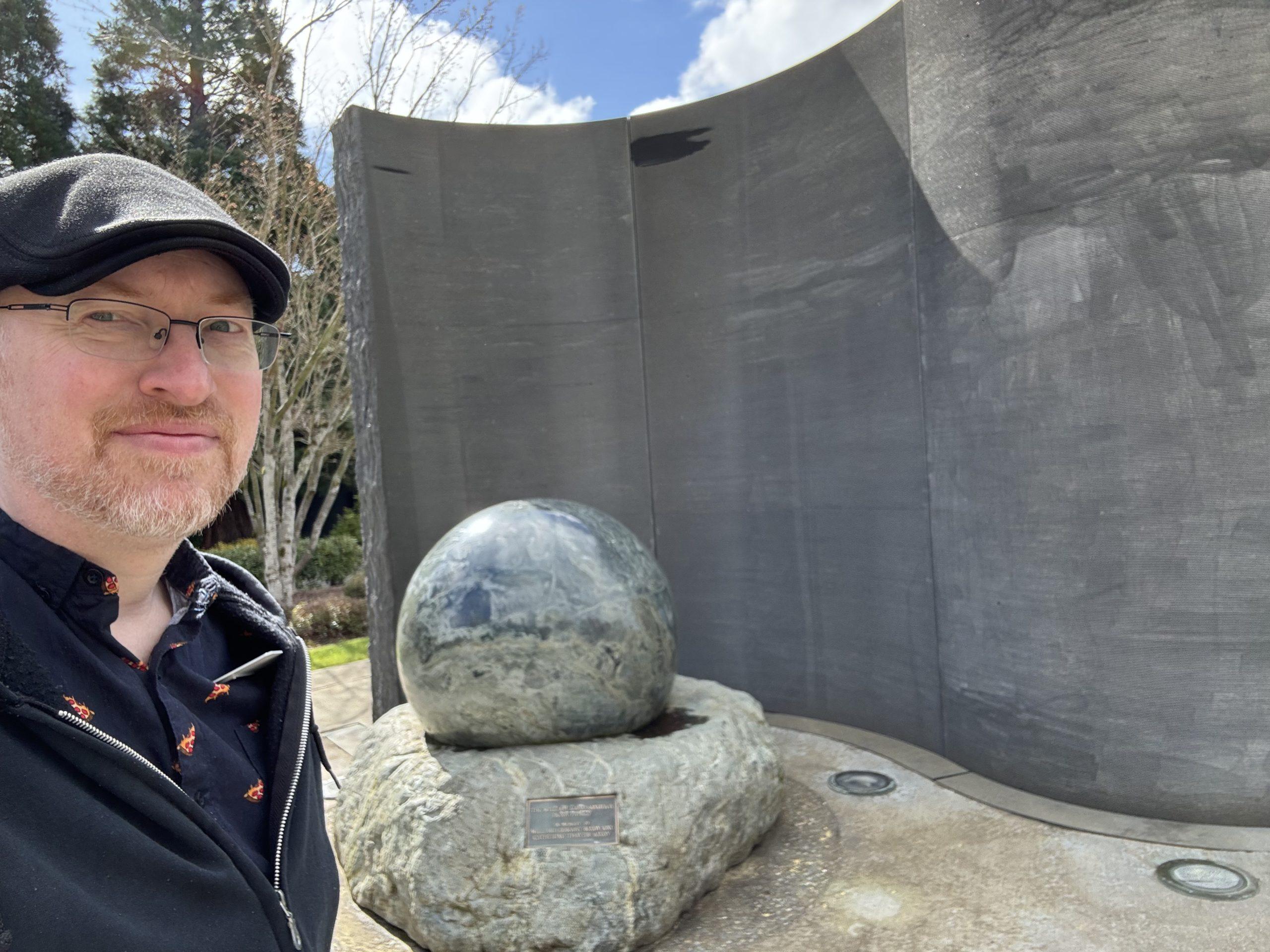 Me in front of an outdoor sculpture of a large spherical rock seated in a depression in another rock in front of a curved, textured grey wall.