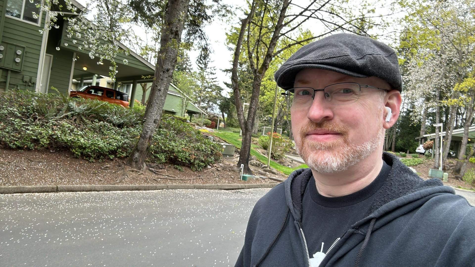 Me outside in our neighborhood, in front of trees, bushes, and green-painted buildings, wearing a t-shirt, hoodie, hat, and with Apple AirPods in my ears.