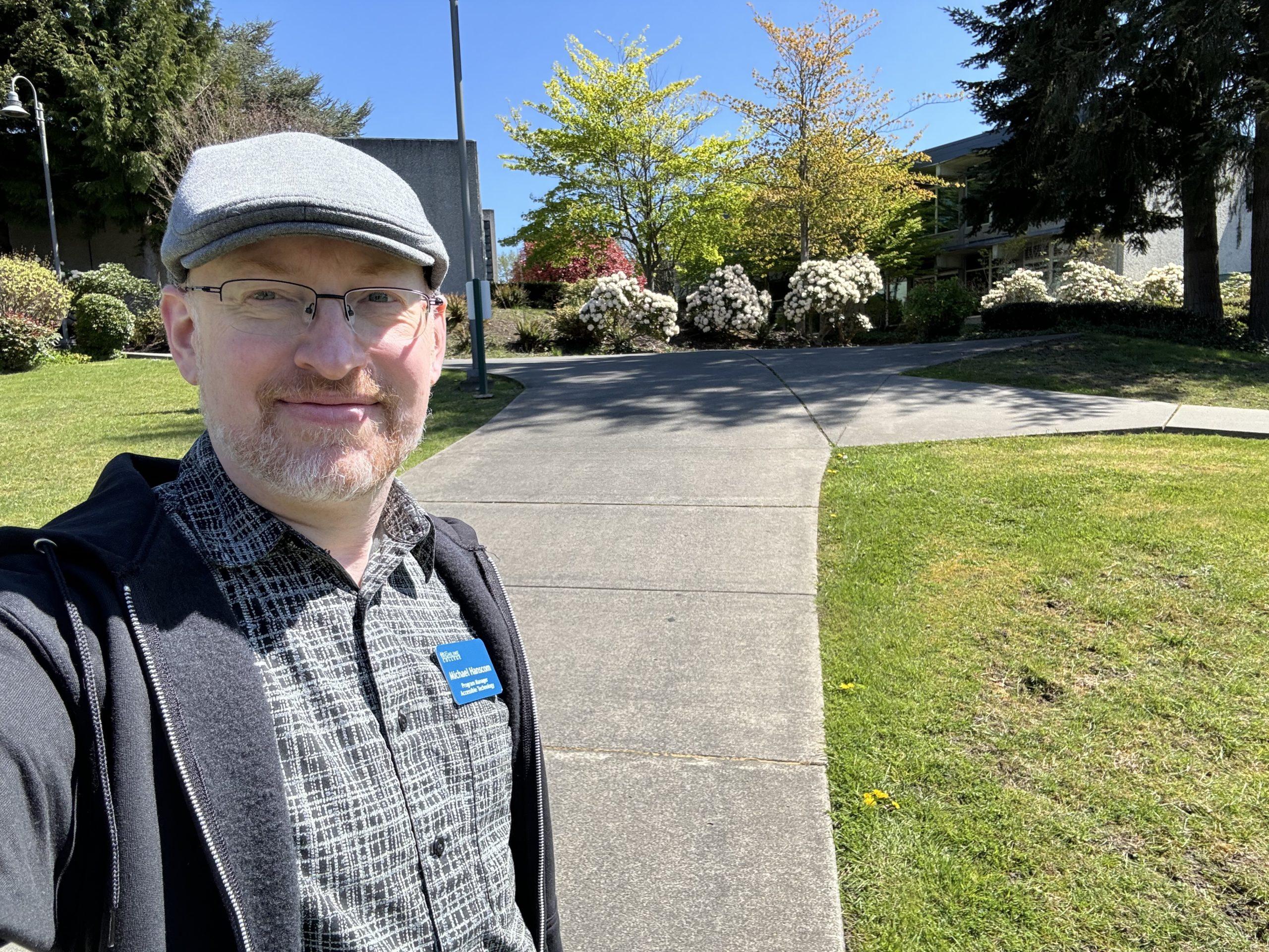 Me outside in the sun on a concrete path between two grassy areas, with bright green trees and flowering bushes in the background.