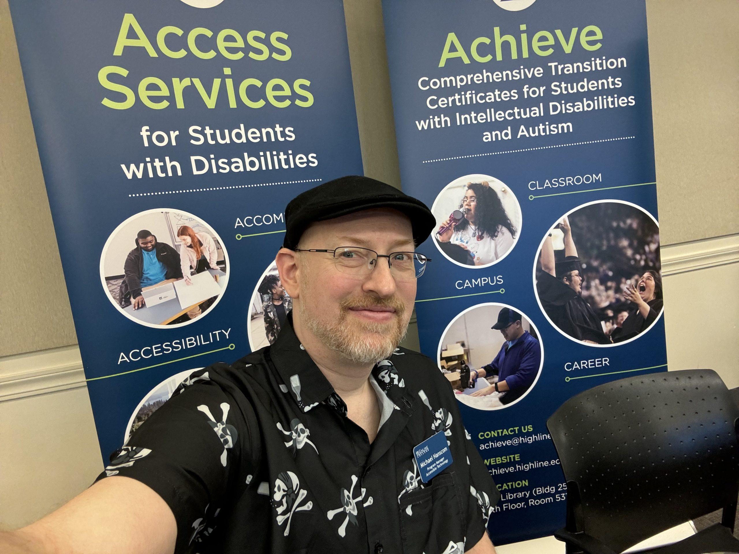 Me wearing a black short-sleeve button-up shirt with a skull and crossbones pattern, in front of signs advertising the Access Services department for students with disabilities and the Achieve program for students with intellectual disabilities.