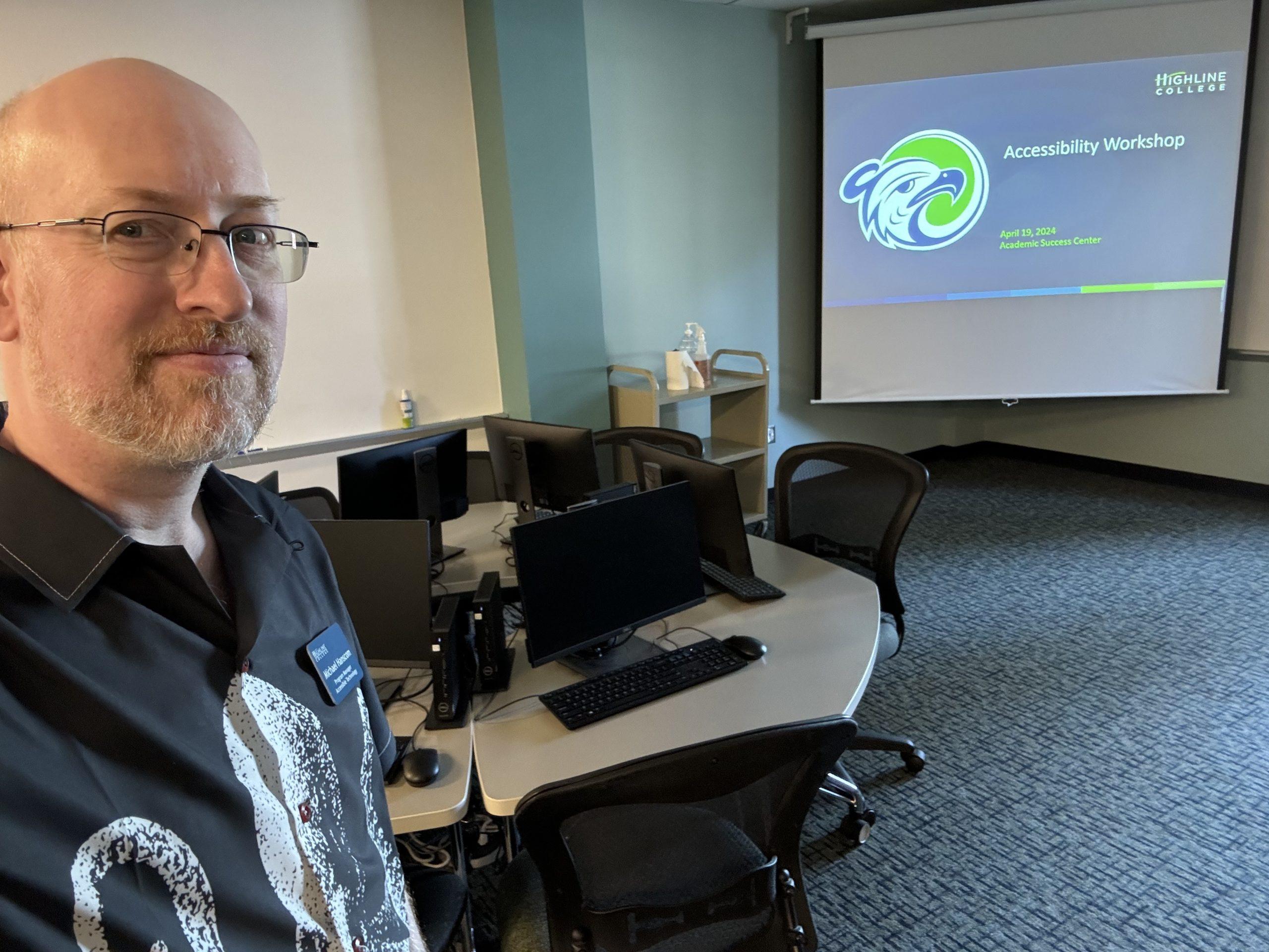 Me in a computer lab classroom, in front of a round table with several inactive workstations. Against the wall is a screen showing a slide with the Highline College logo and the title 'Accessibility Workshop'.