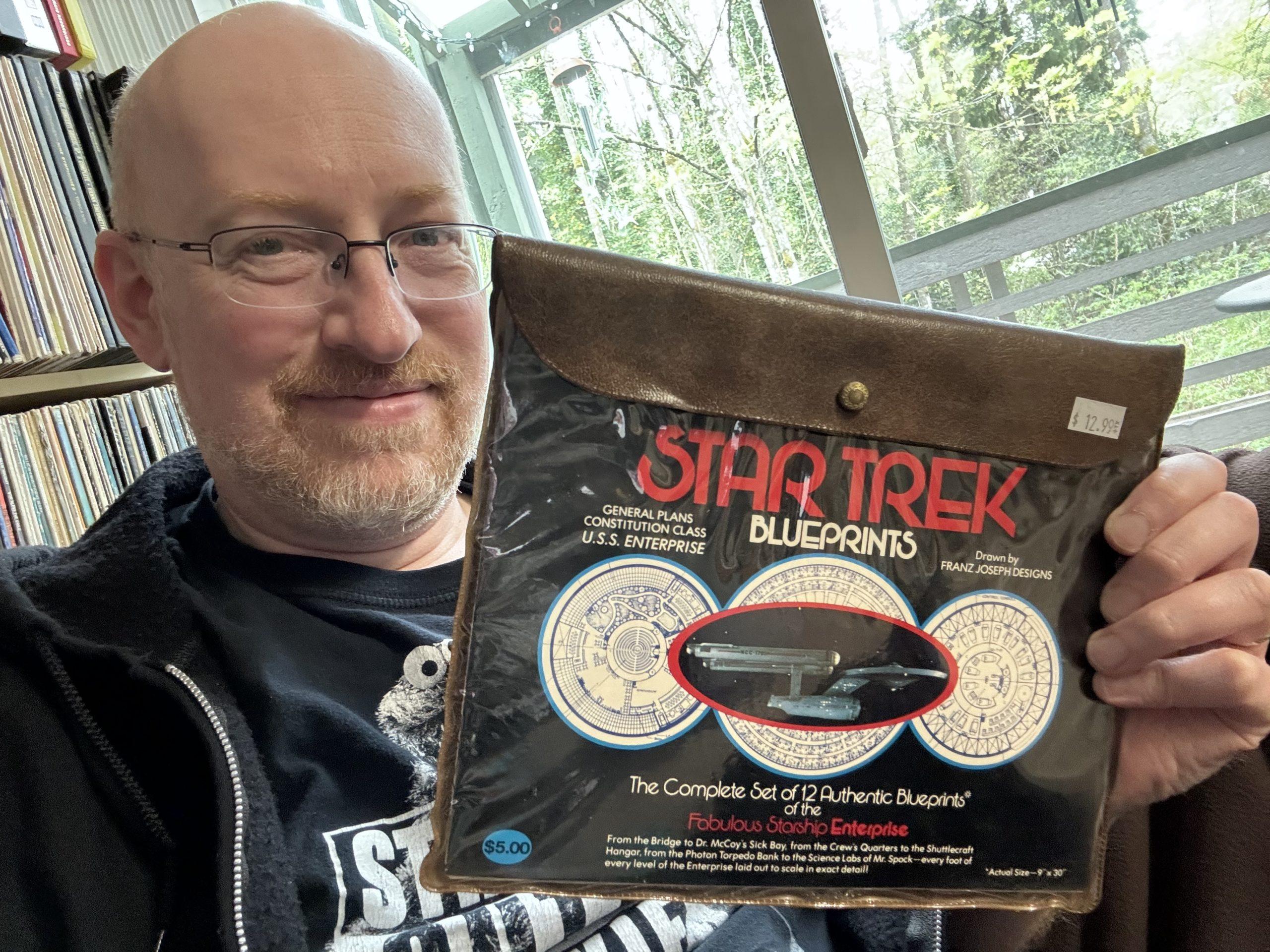 Me holding a brown faux-leather pouch with a clear front, which contains the classic Star Trek blueprints of the original series USS Enterprise.