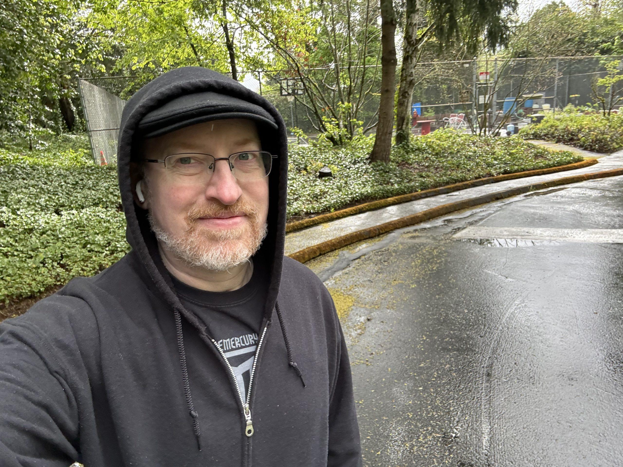 Me, wearing hat and hoodie zipped up and with the hood pulled up over my head, outside on wet pavement in front of green bushes and trees by a fenced-in tennis court.