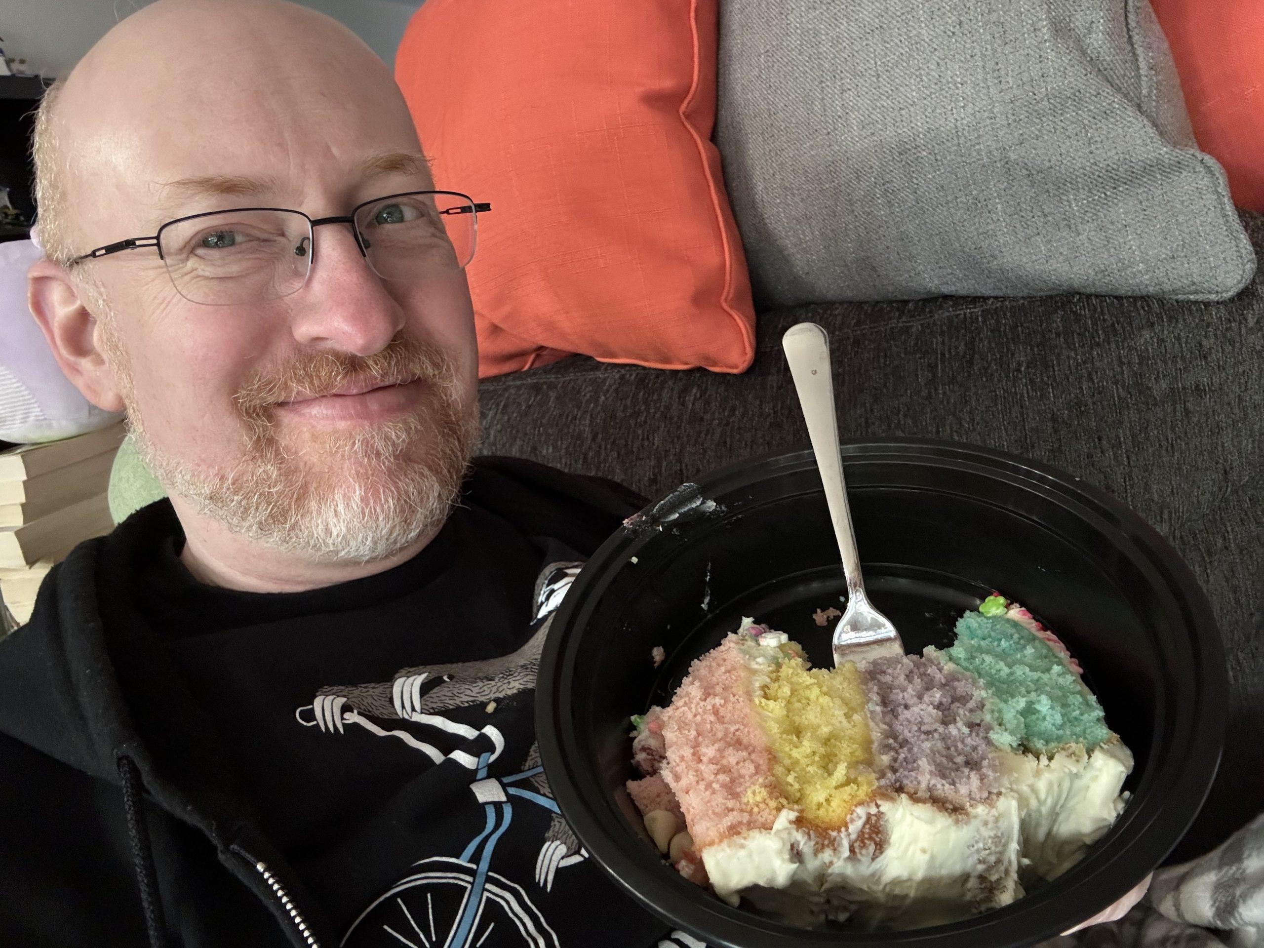 Me on our couch, holding a plate with a piece of cake with four layers, one pink, one yellow, one purple, and one green.