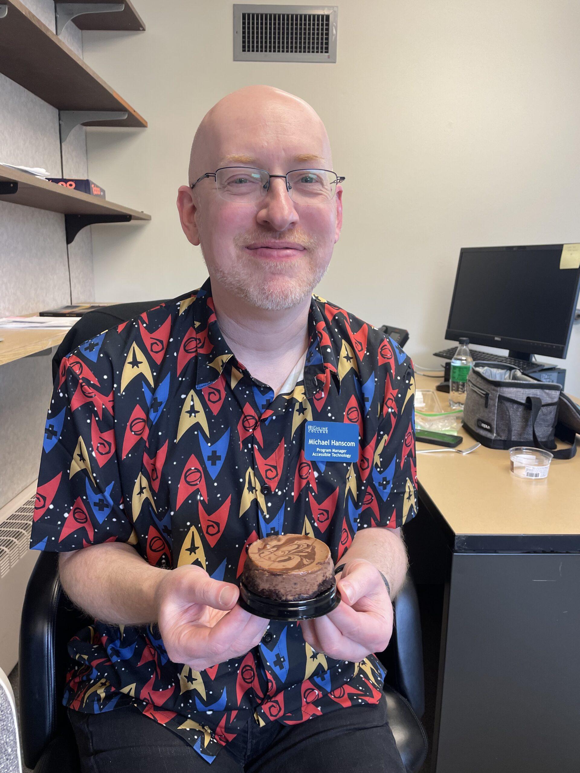 Me sitting at an office desk holding a small chocolate cheesecake.