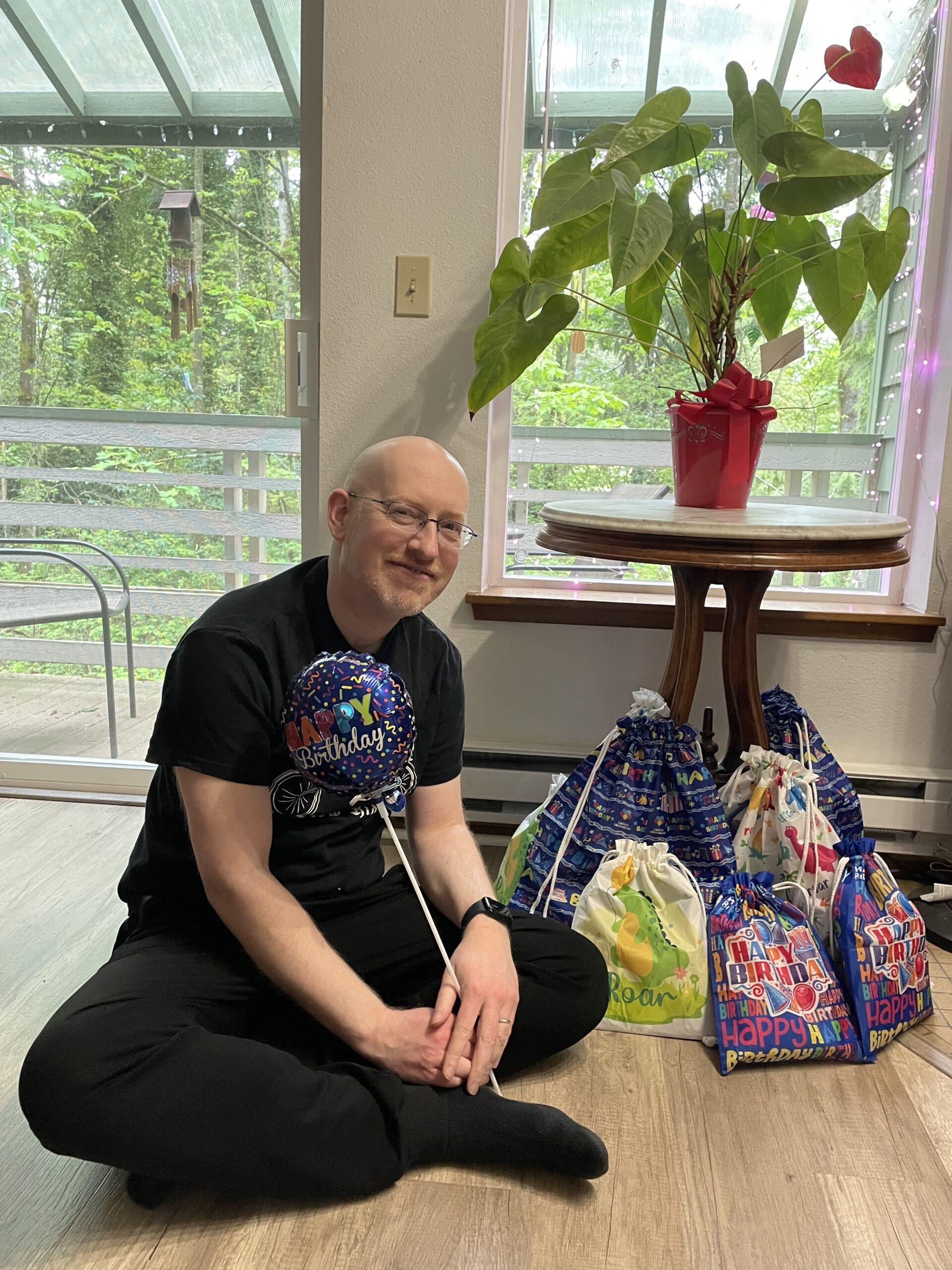 Me sitting on our living room floor, holding a 'Happy Birthday' balloon, in front of a small stack of colorfully bagged birthday presents.