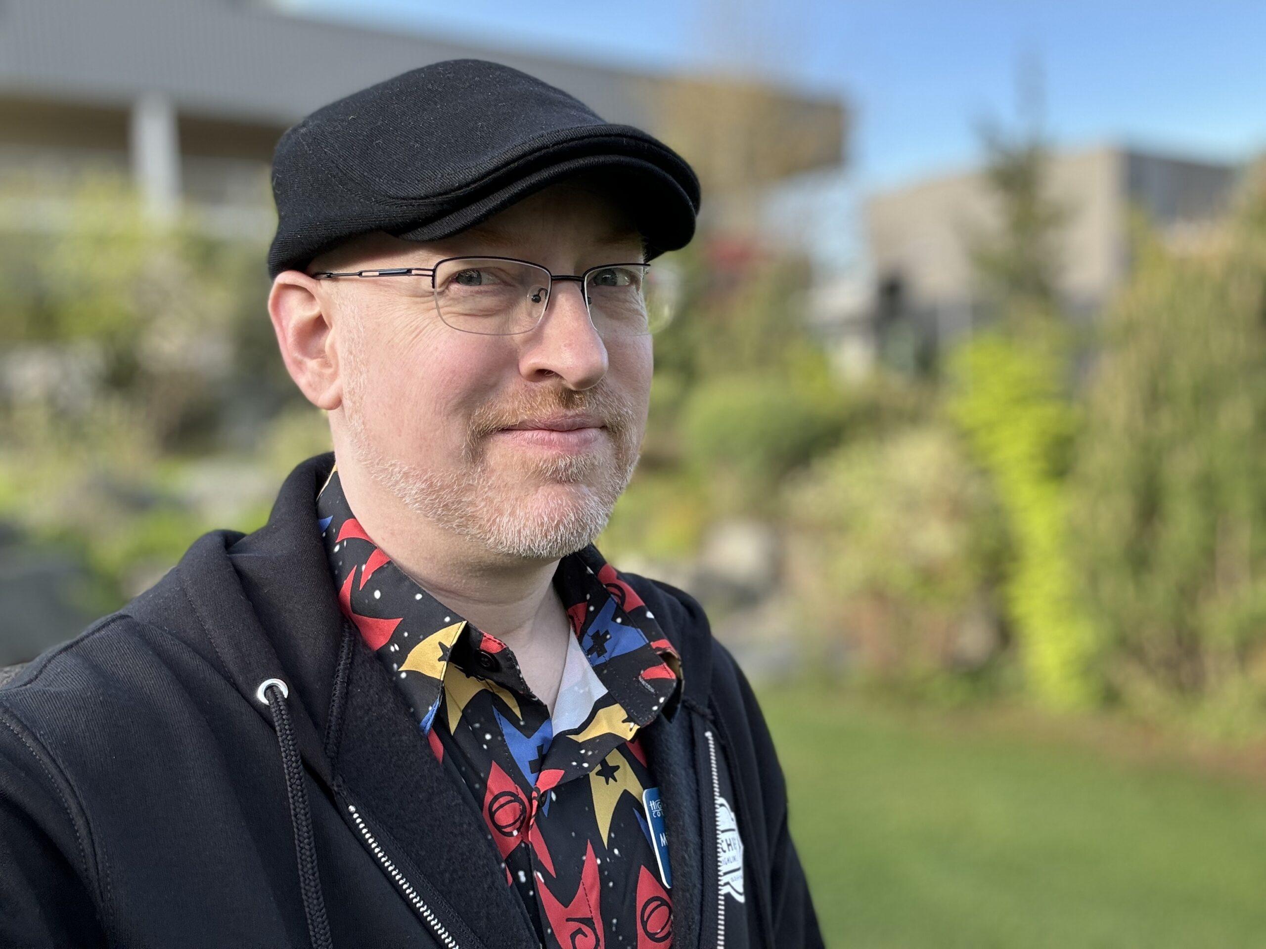 Me outside in front of blurred grass and trees, wearing a black cap, black hoodie, and button-up shirt with a pattern of Star Trek division logos. I'm a white man with glasses and neatly trimmed greying red beard.