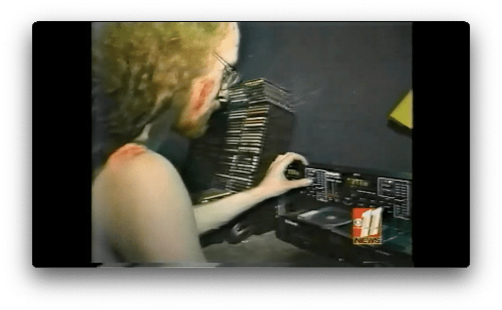 A somewhat low-quality screengrab of a video clip of me DJing in 1998. The shot is from behind me over my right shoulder. I have long curly red hair dyed black at the ends. I'm not wearing a shirt, and I have red streaks of mud on my cheek and shoulder. I have one hand on the tempo control of a Numark CD DJ mixer. There is a rack of CDs to my left.