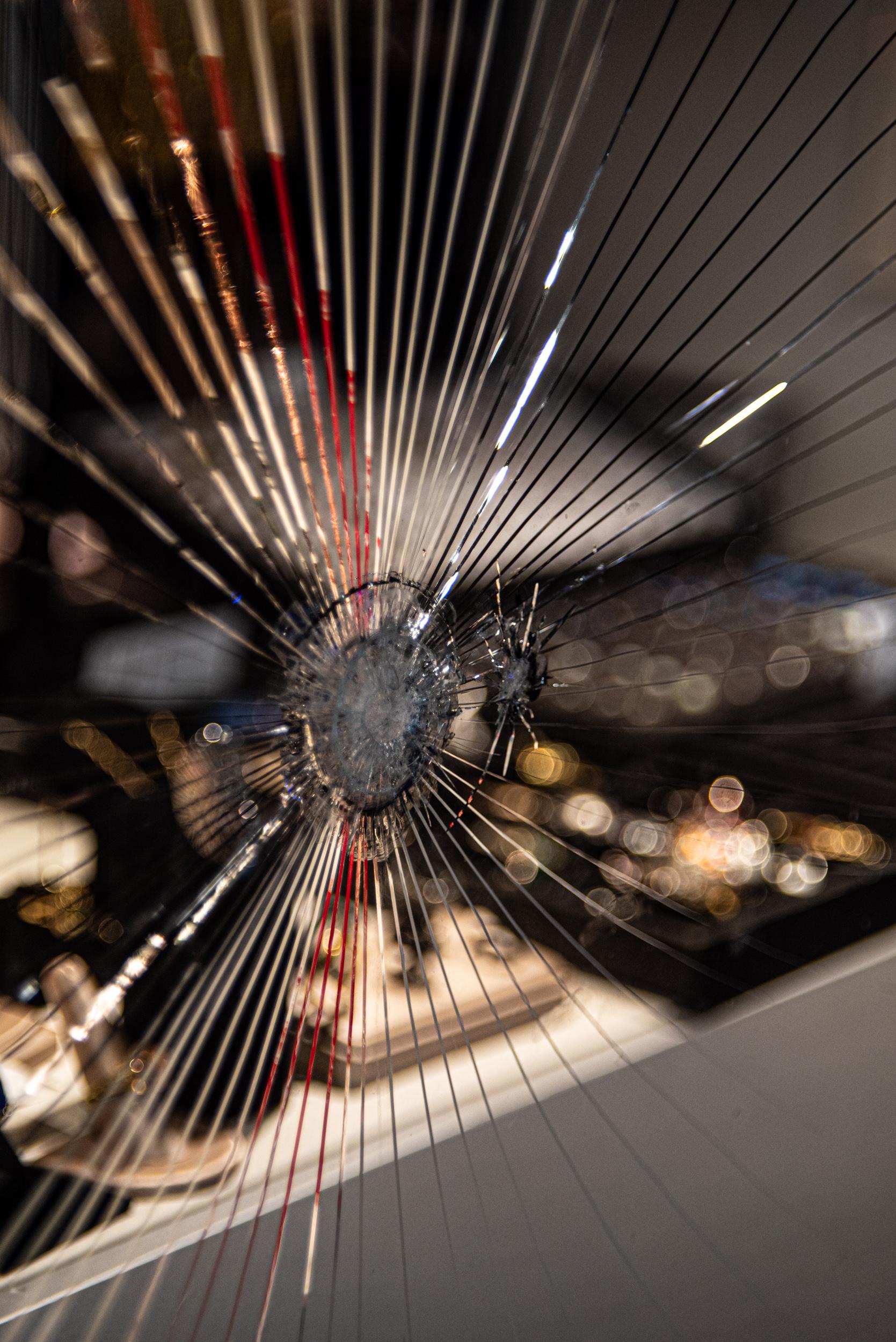 A pane of shattered glass, with cracks radiating out from a central impact point; a smaller impact point is just to one side. Behind the glass and artfully out of focus are sparkling pieces of jewelry.