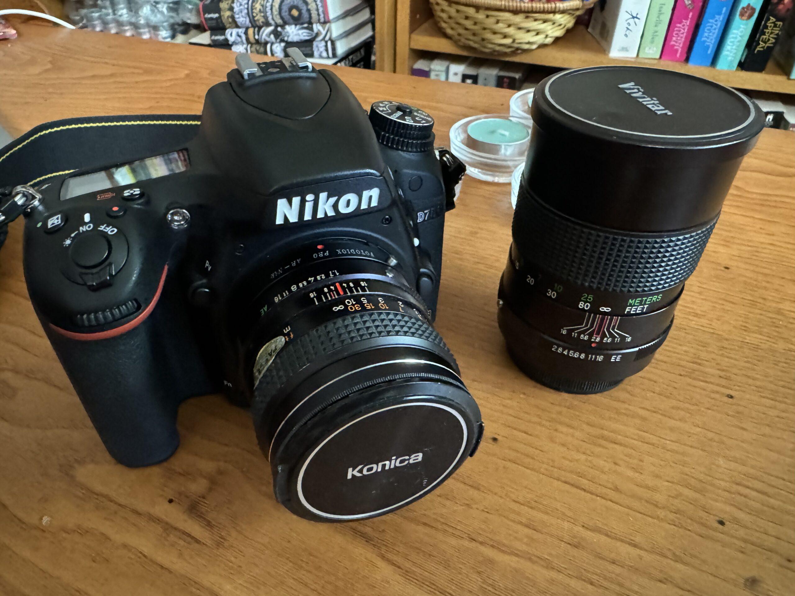 A Nikon D750 DSLR sits on a coffee table. Attached to it is an 1970s Konica lens, while a Vivitar lens from the same era sits nearby.