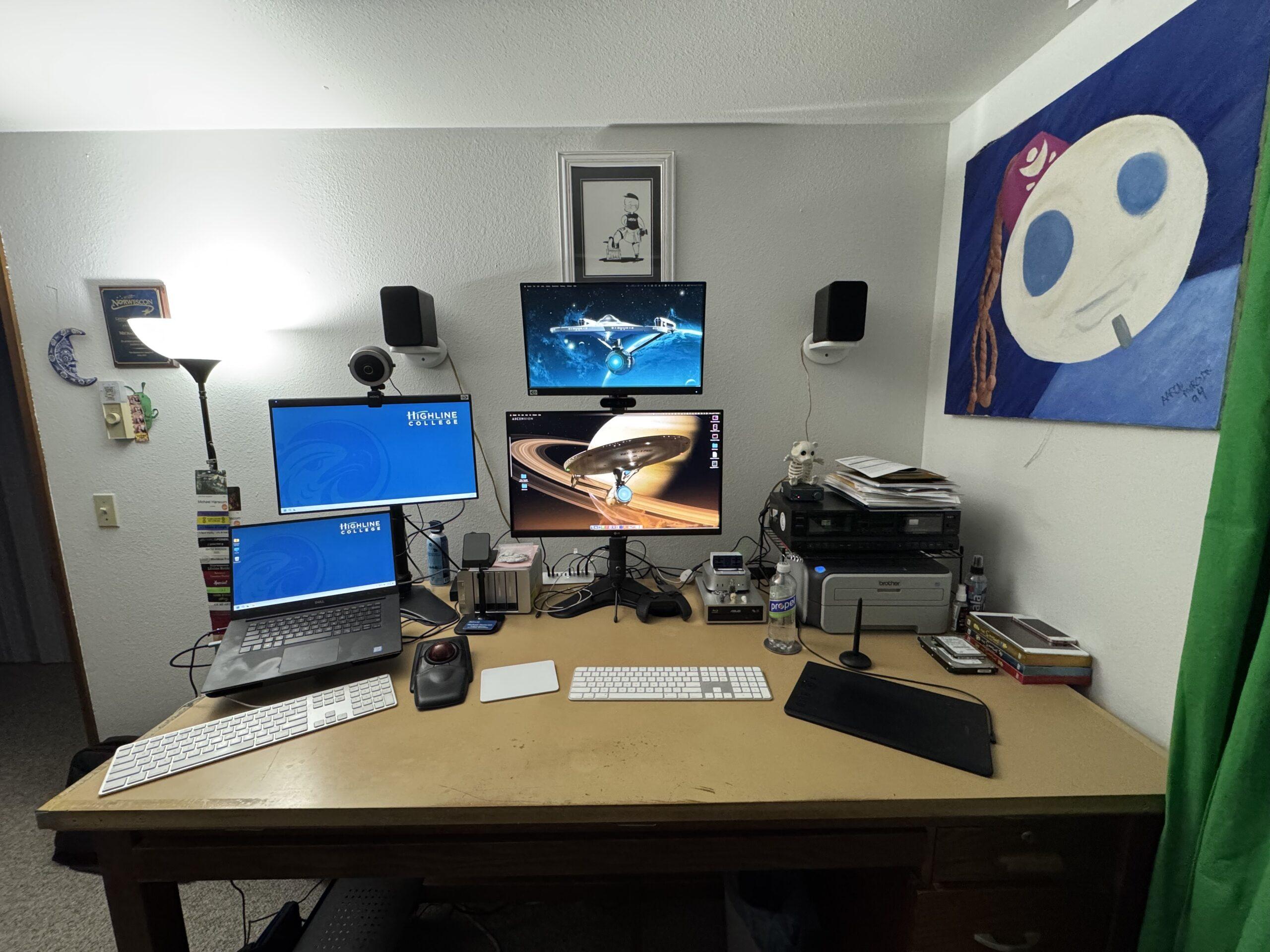 A large with a PC laptop with an external monitor and a Mac Mini with two external monitors. There are two keyboards, one trackball, one track pad, and one drawing tablet towards the front of the desk. A printer sits on the right side of the desk. On the walls behind the desk are speakers and artwork, including a black-and-white cartoon sketch and a white-and-blue painting of a figure with a round head and two large blue circles for eyes, wearing a red fez.